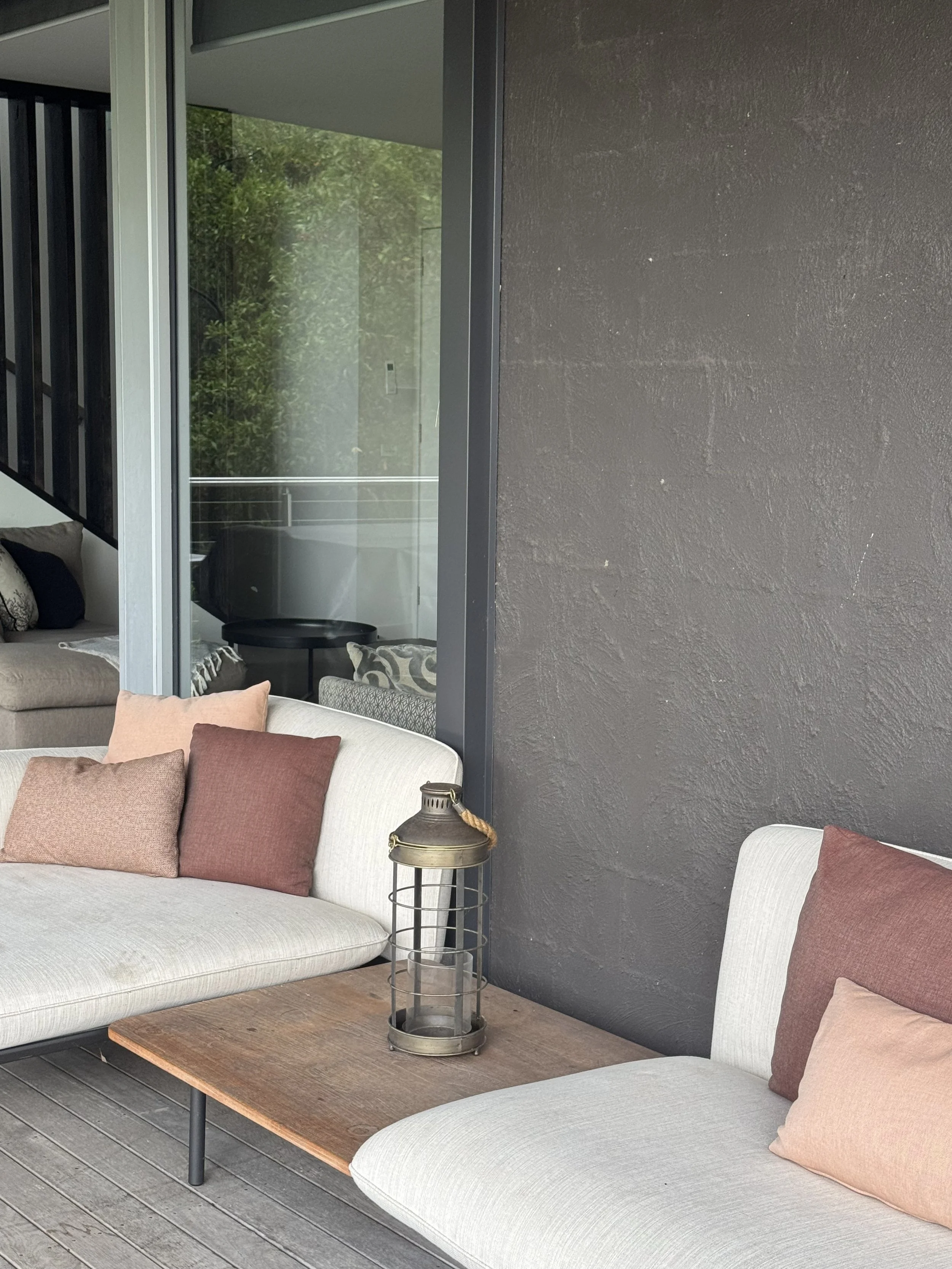 Outdoor patio seating with two beige sofas with pink and red pillows, a wooden coffee table, and a lantern, with a view of a house interior through sliding glass doors.