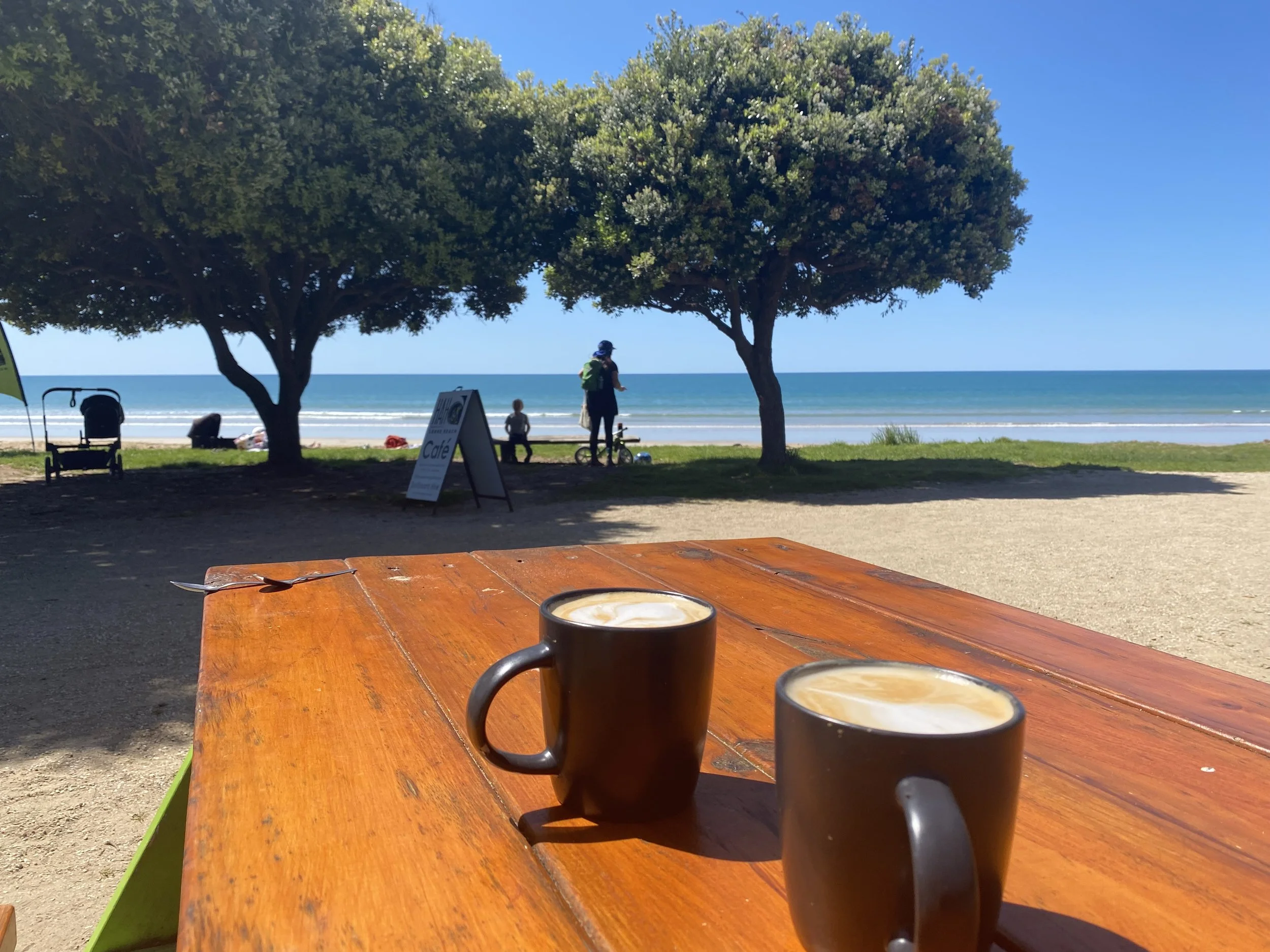 Two black coffee mugs with foam on top on a wooden picnic table near the beach with trees, sand, ocean, and people in the background.