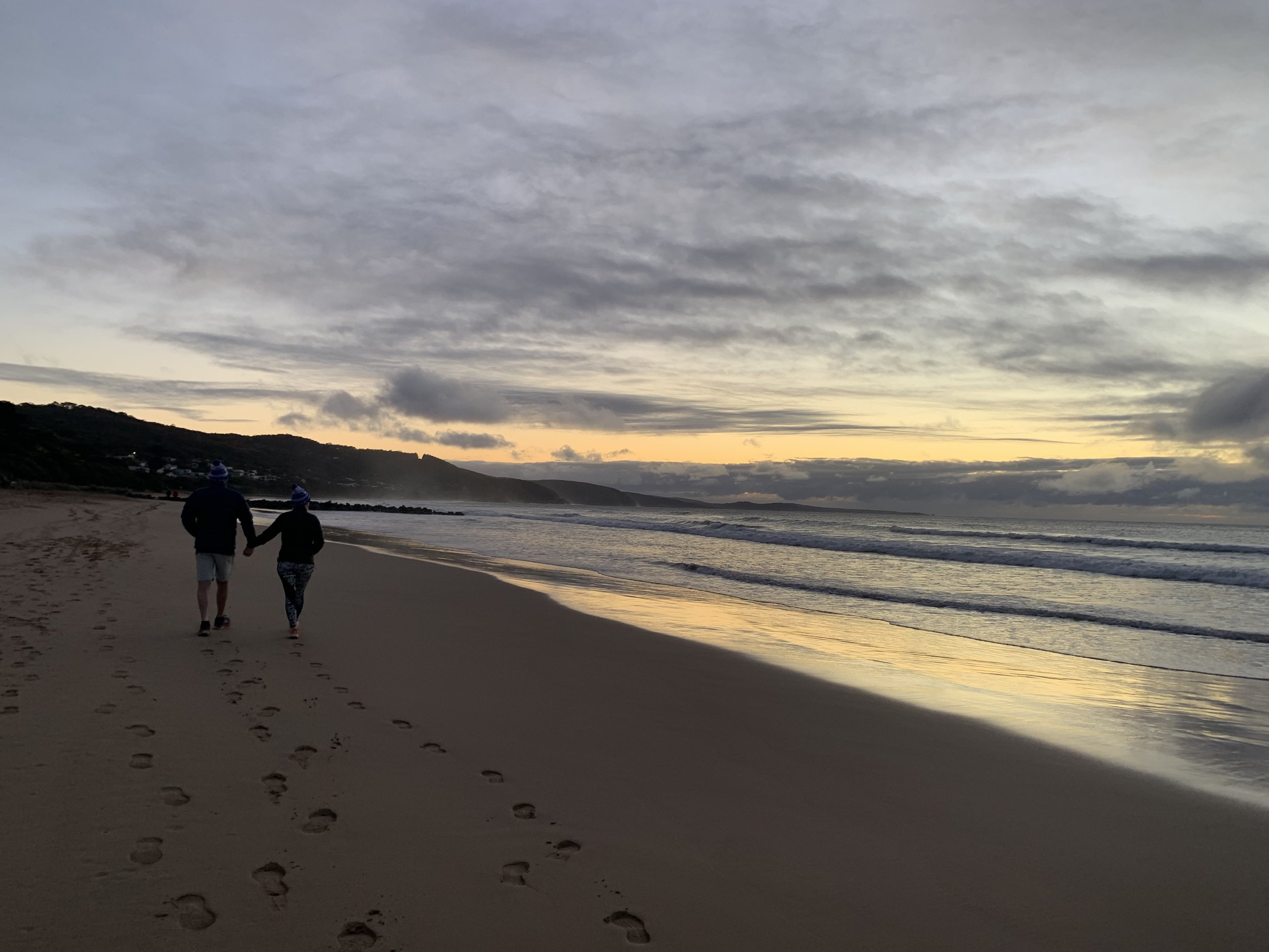 A couple walking hand in hand along a sandy beach during sunset, with footprints in the sand and cloudy sky.