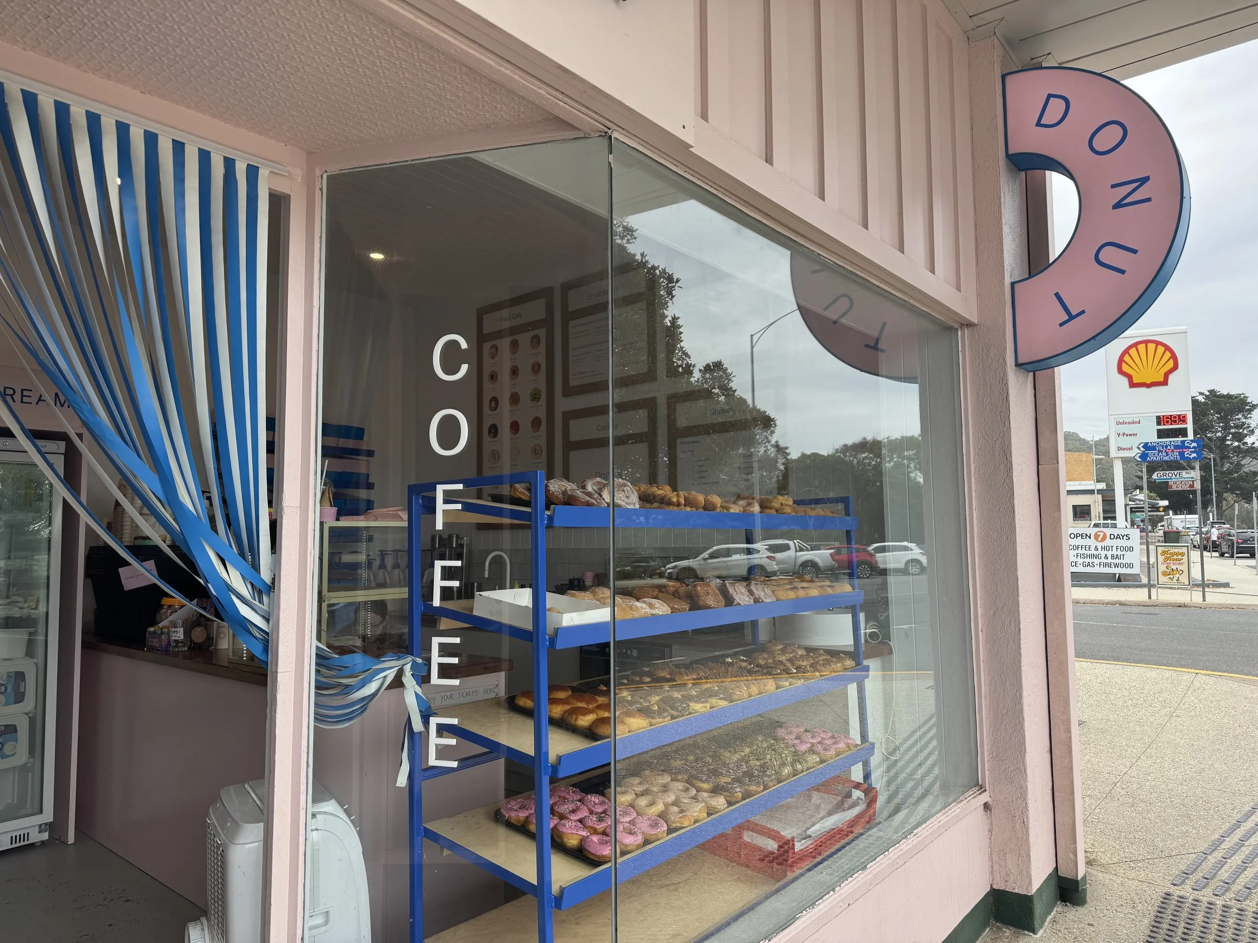 Bakery storefront window displaying baked goods on blue racks, with a pink and blue donut-shaped sign reading 'DONUT' outside, and a Shell gas station sign in the background.