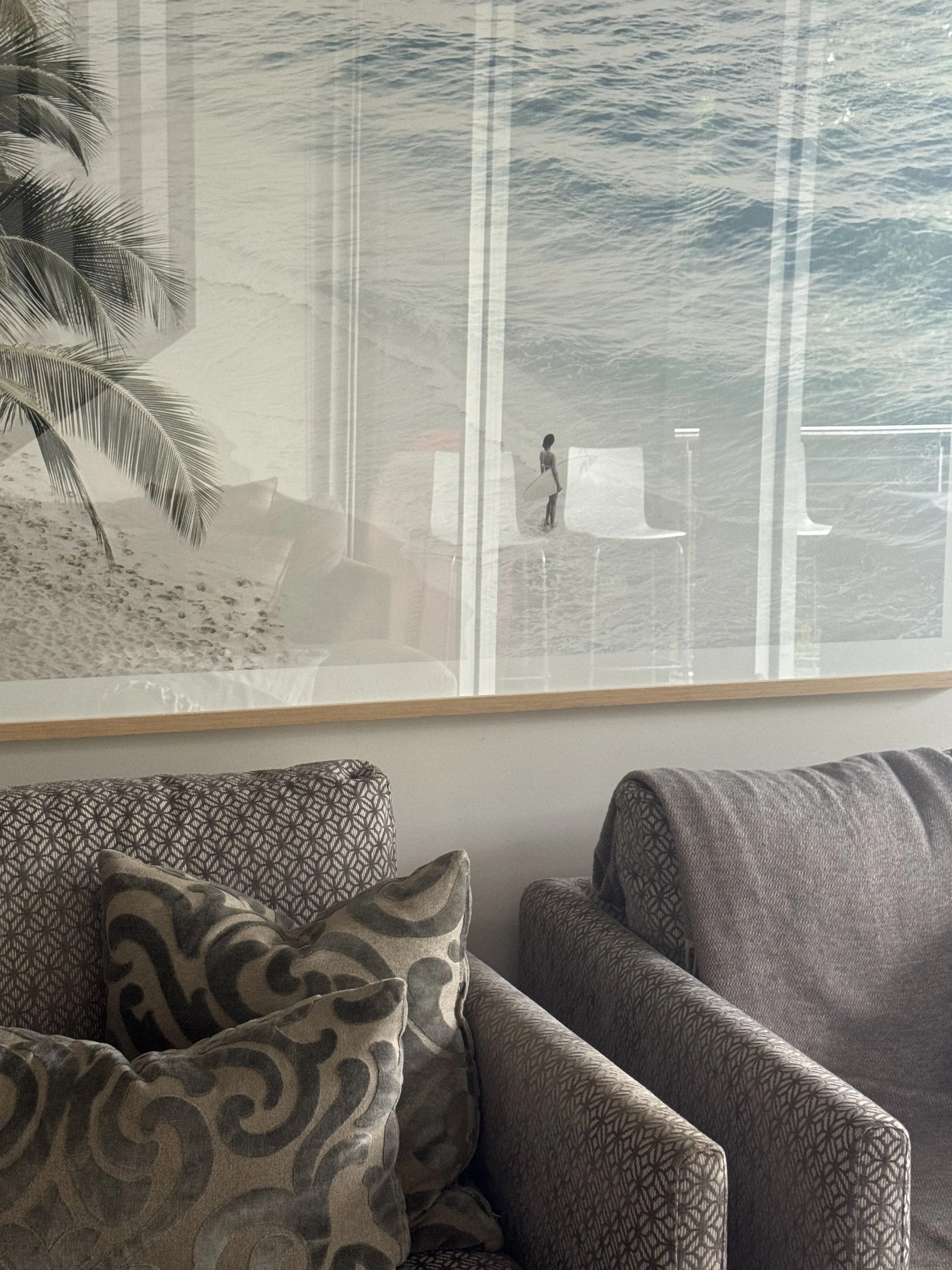 View of a beach scene with a woman holding a surfboard, seen through a window from inside a room with patterned sofa cushions.