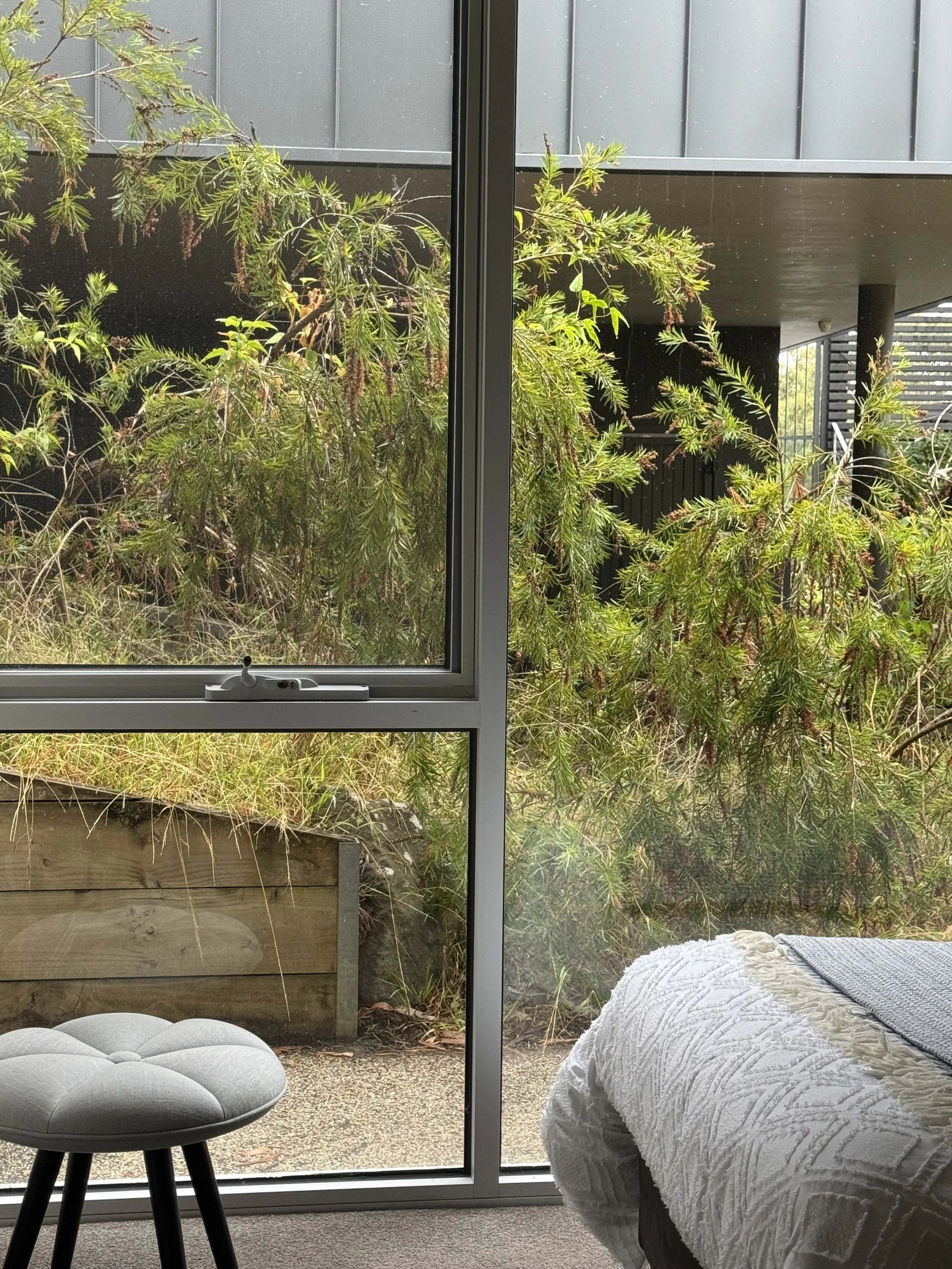 View through large glass window showing outdoor greenery, including a large bush and a wooden planter, with part of a bed and a small cushioned stool inside.
