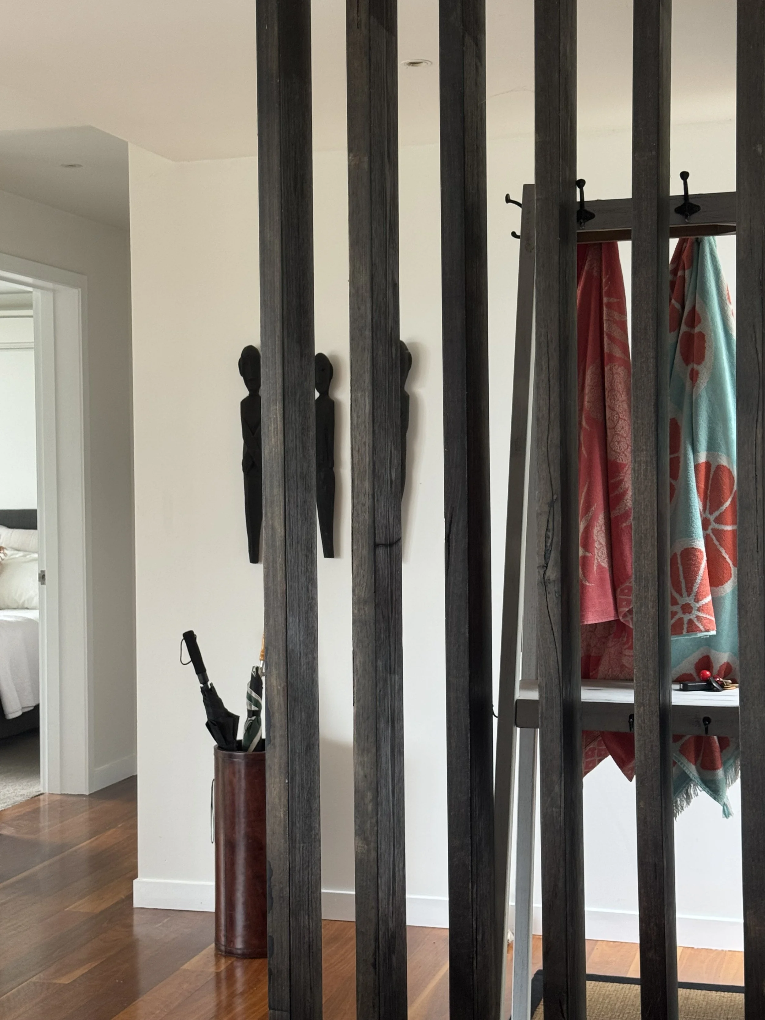Part of a living space seen through black wooden vertical slats in an interior divider. Visible are a white wall with three black tribal-style wall sculptures, a brown umbrella stand with umbrellas, and a room with a bed. On the right side, there is 