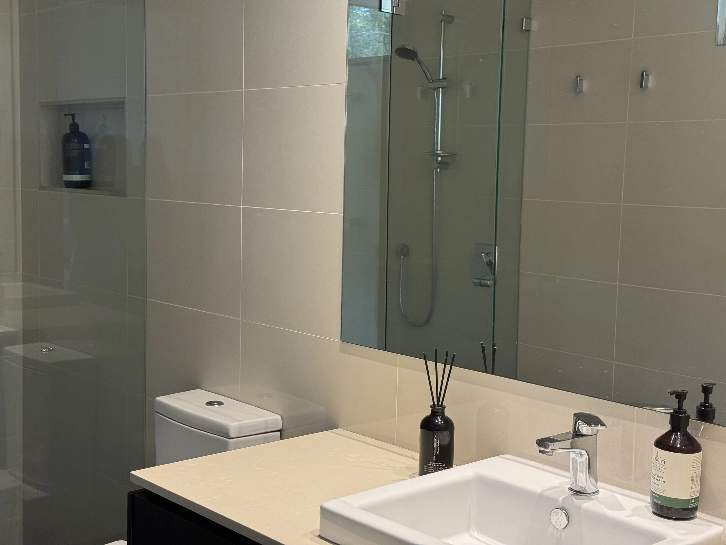 Modern bathroom with large mirror, beige tiles, and a white sink with black countertop, black soap dispenser, diffuser, and a shower area with a glass partition and a built-in shelf with a bottle.
