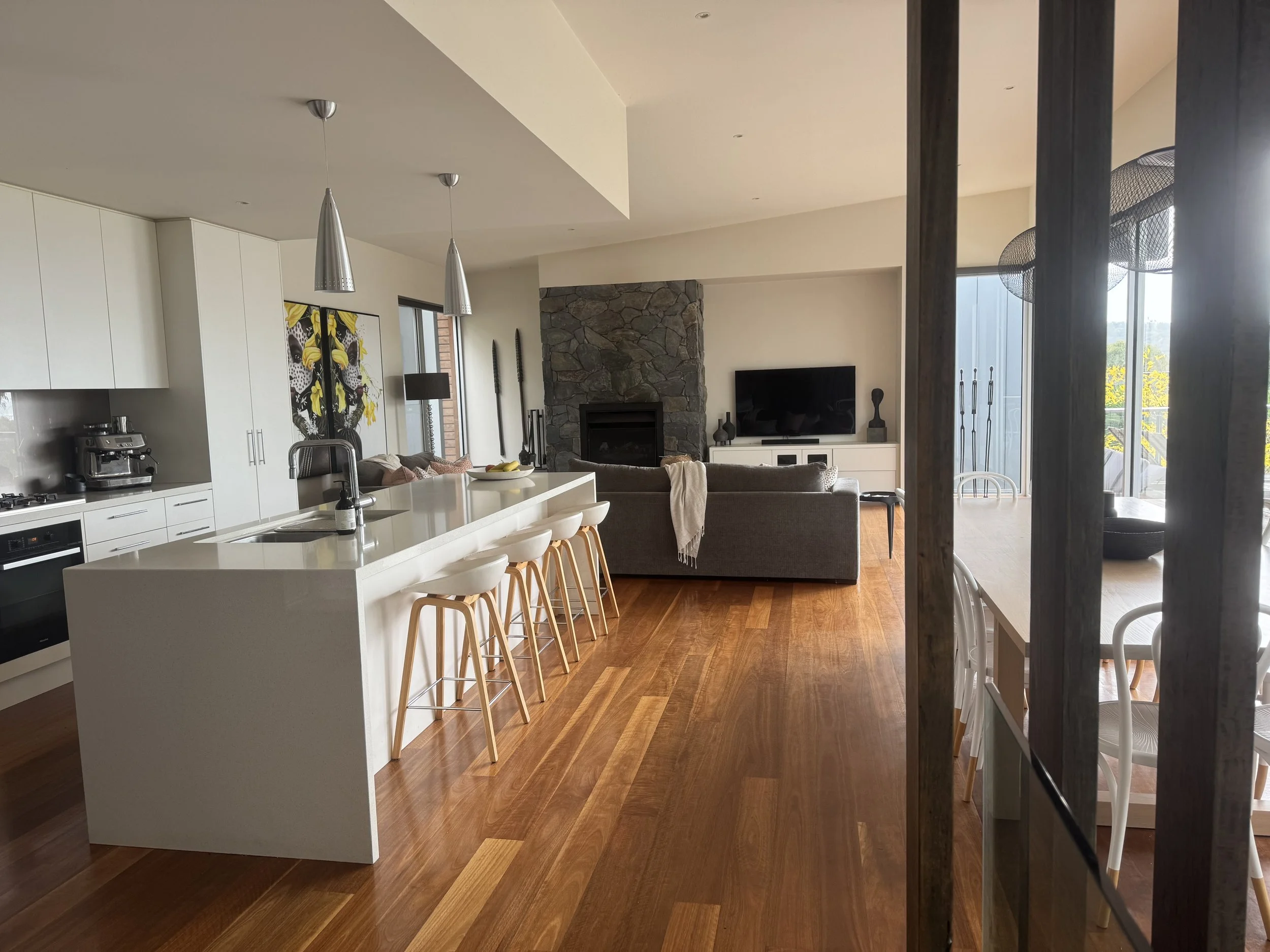 Open-plan living space with a kitchen island, white cabinets, and sitting area with a stone fireplace, flat-screen TV, and chairs near large windows.