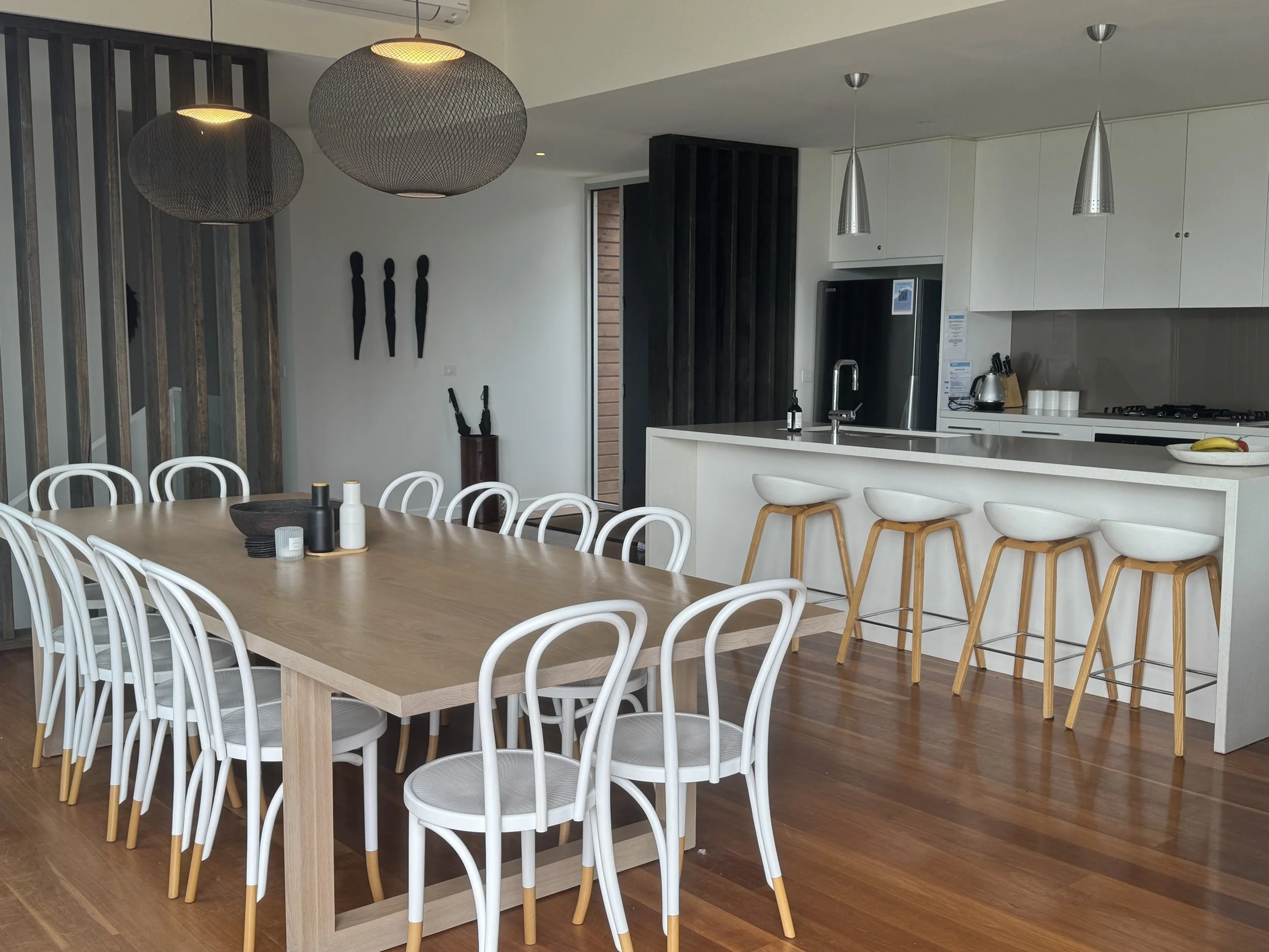 Modern kitchen and dining area with a large wooden table, white chairs with wooden legs, a kitchen island with three white barstools, and pendant lights.