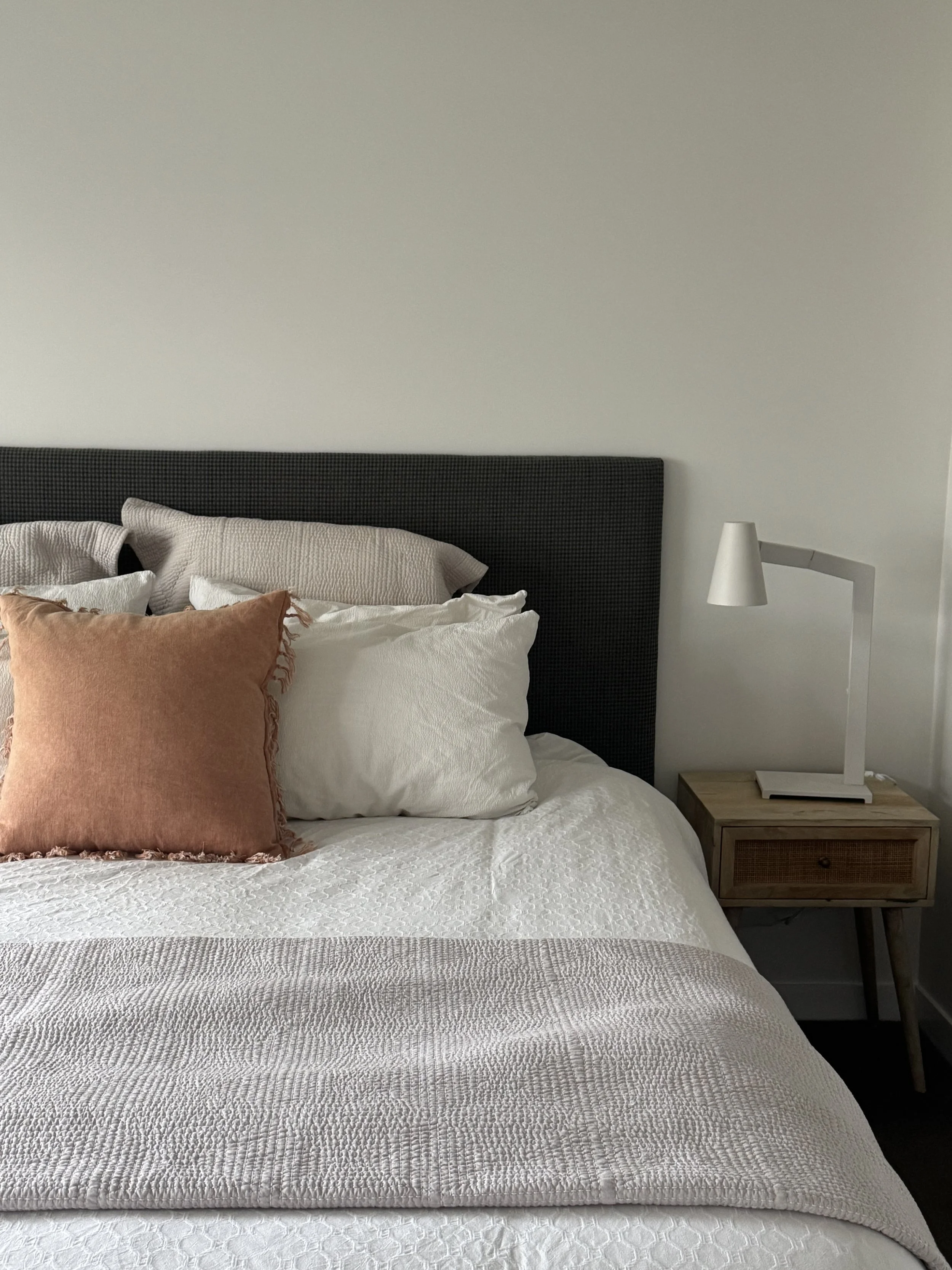 A neatly made bed with white quilt, beige pillows, and a rust-colored decorative pillow, next to a modern white nightstand with a white lamp and a closed book, against a plain white wall.