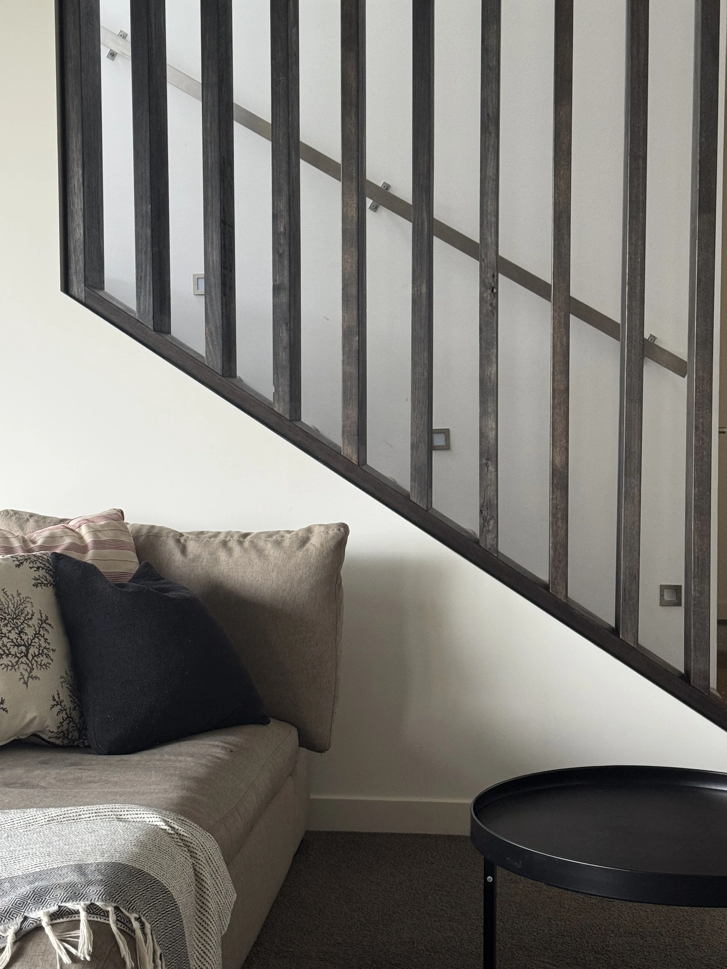 Living room corner with beige sofa, black and patterned decorative pillows, a blank gray throw blanket, a small round black side table, and a wooden staircase railing against a plain white wall.