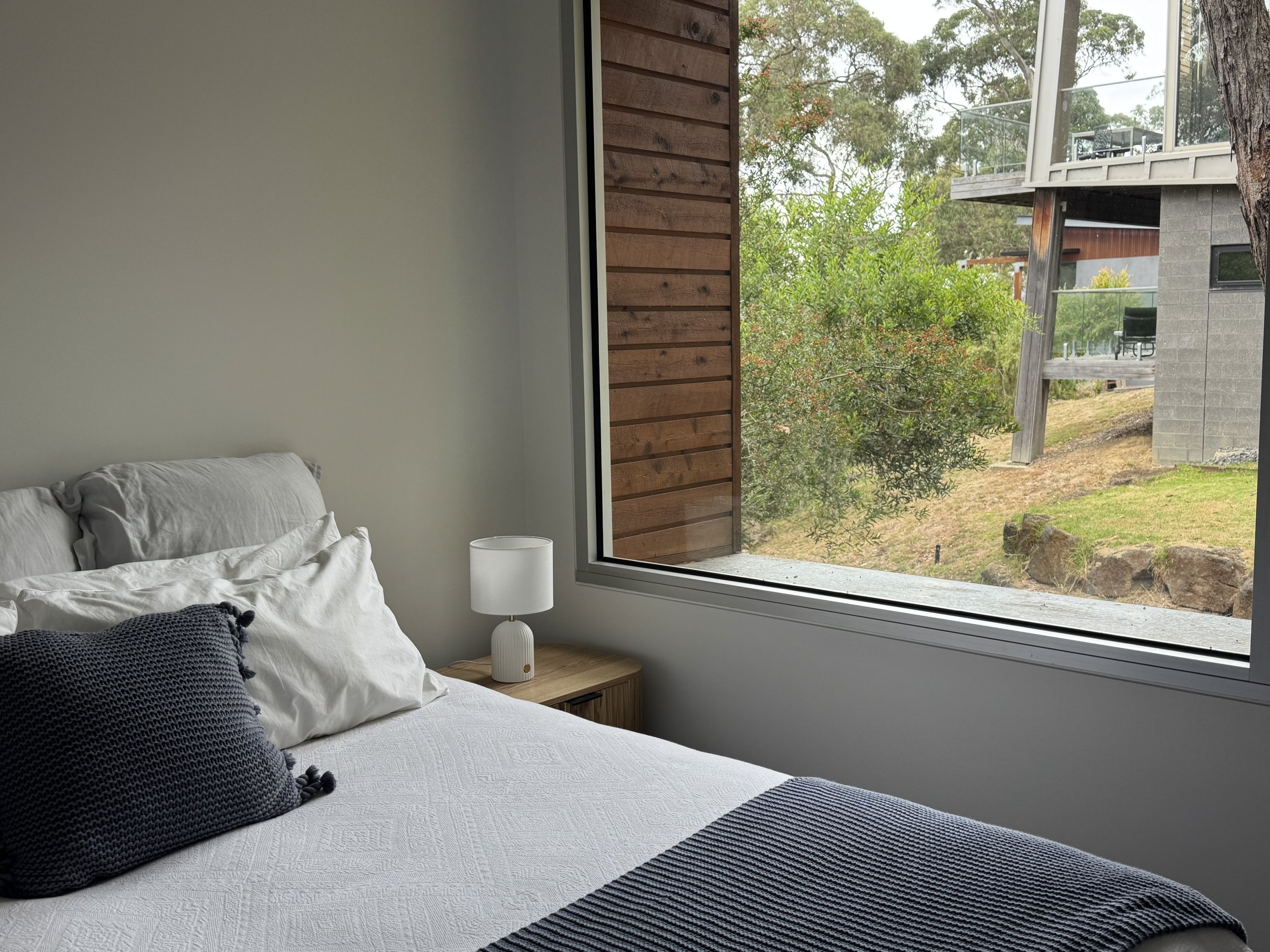 Bedroom with a bed, white bedding, black throw pillow, wooden nightstand with a white lamp, large window showing outdoor greenery and neighboring modern house.