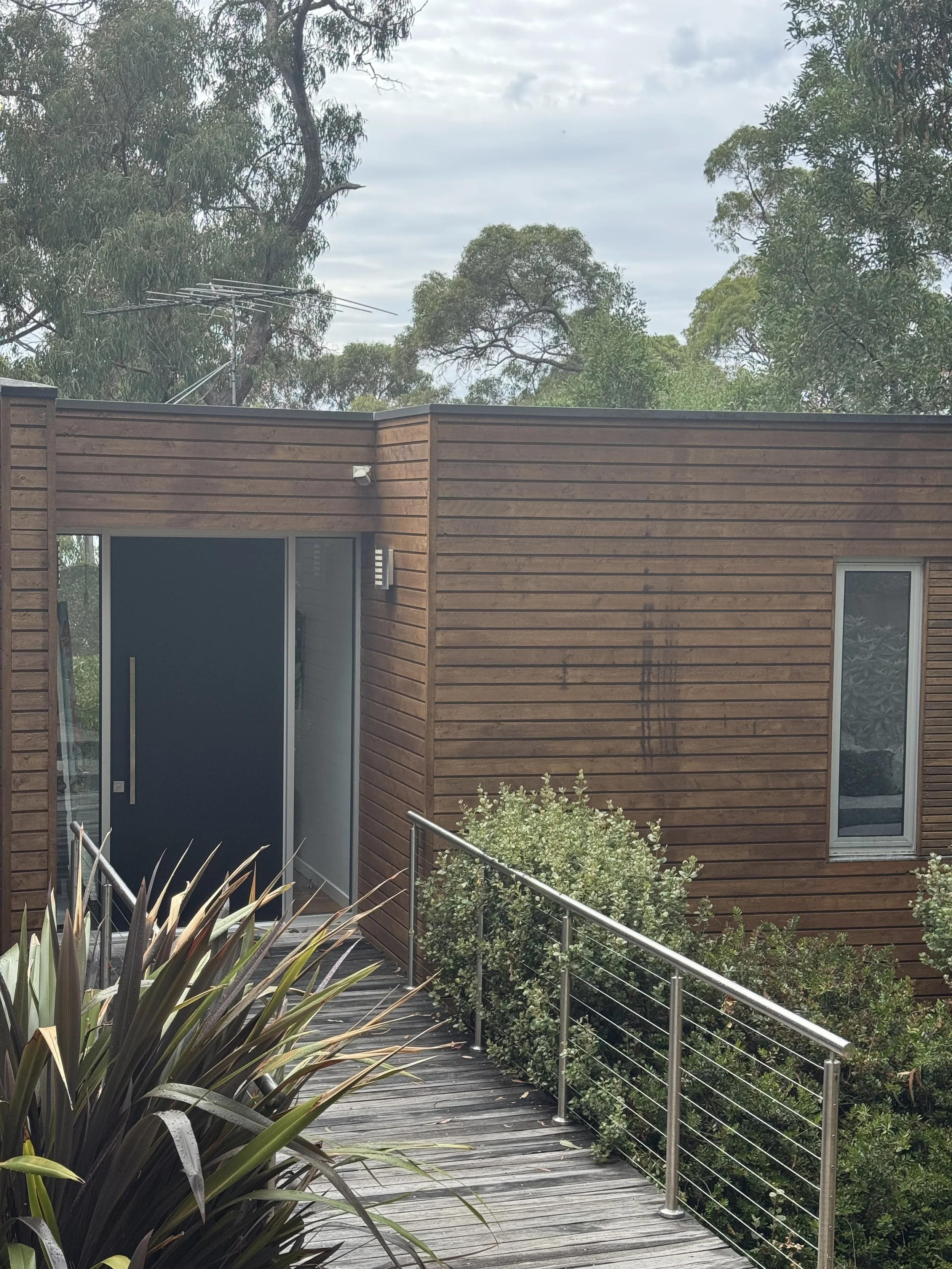 A modern house with wooden exterior siding and a walkway with metal railing, lush green shrubs, and trees in the background.