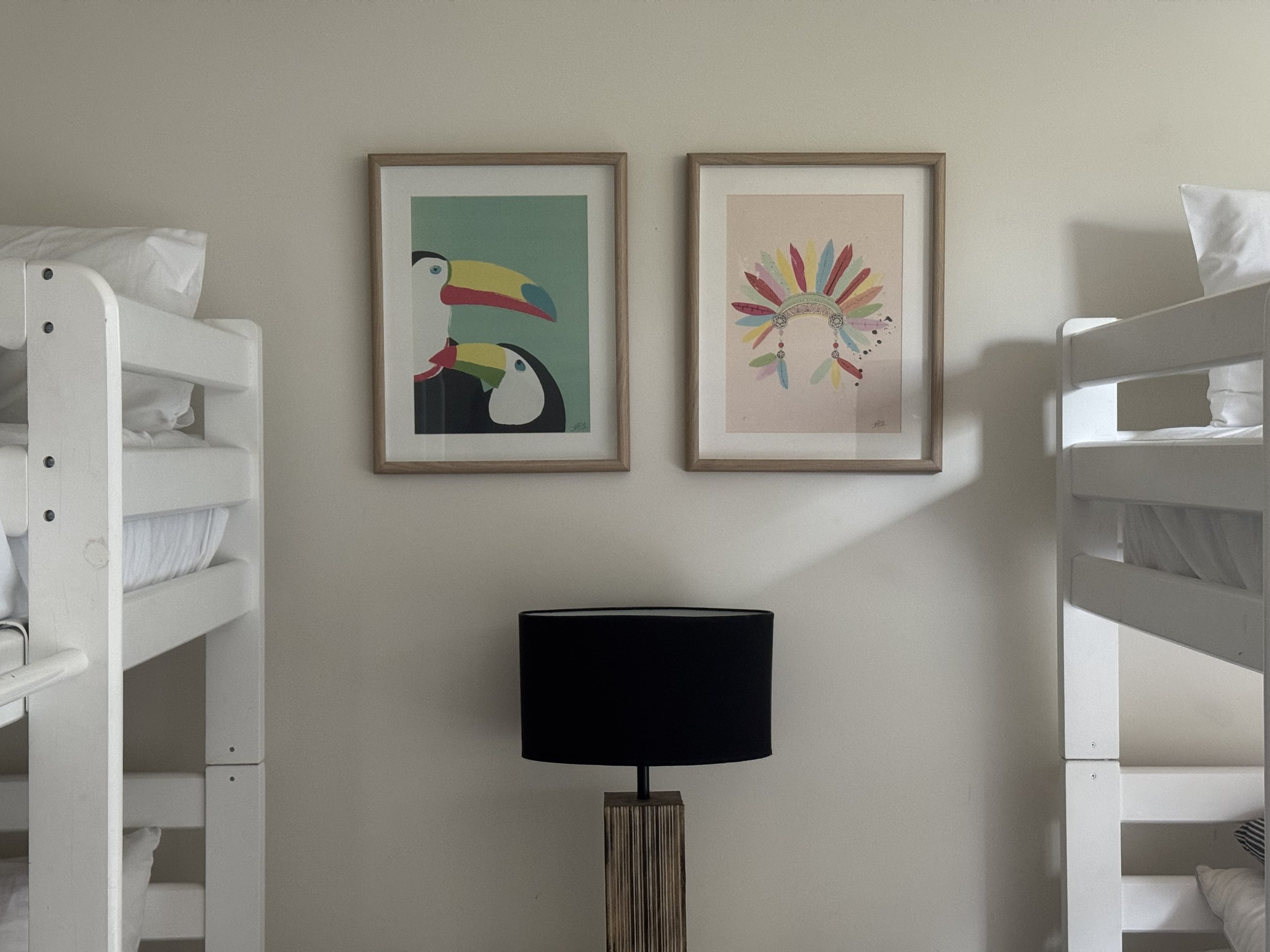 A wall with two framed artwork pieces above a wooden floor lamp with a black lampshade, positioned between two white bunk beds with pillows.