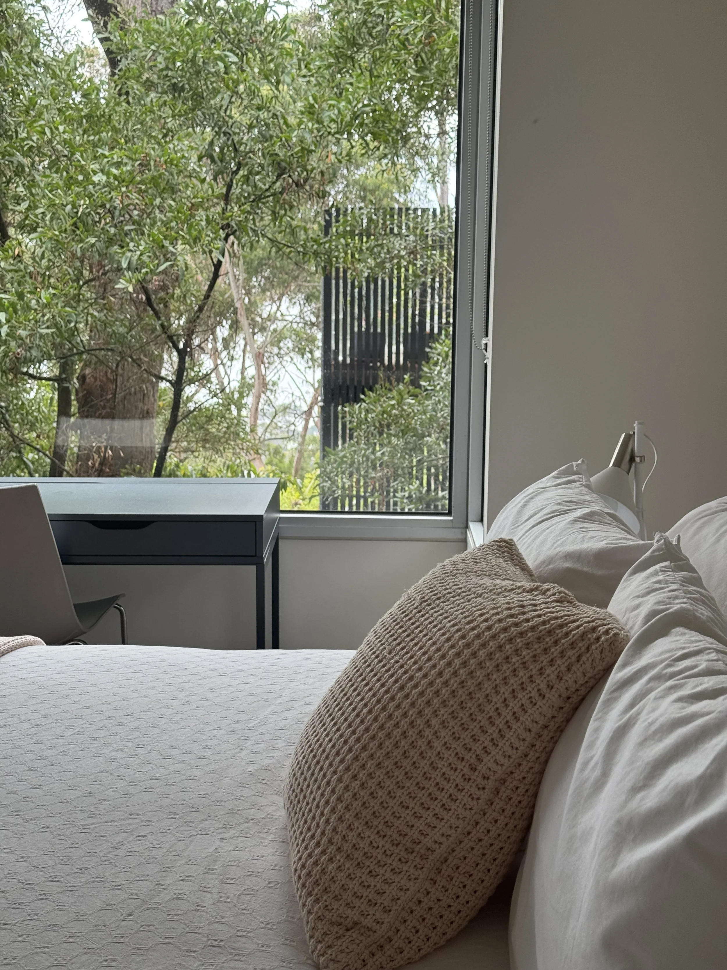 A cozy bedroom with a large bed adorned with white linens and a beige textured pillow. A white bedside lamp is next to the bed. Outside the large window, there are green trees and a black fence, suggesting a garden or outdoor space.