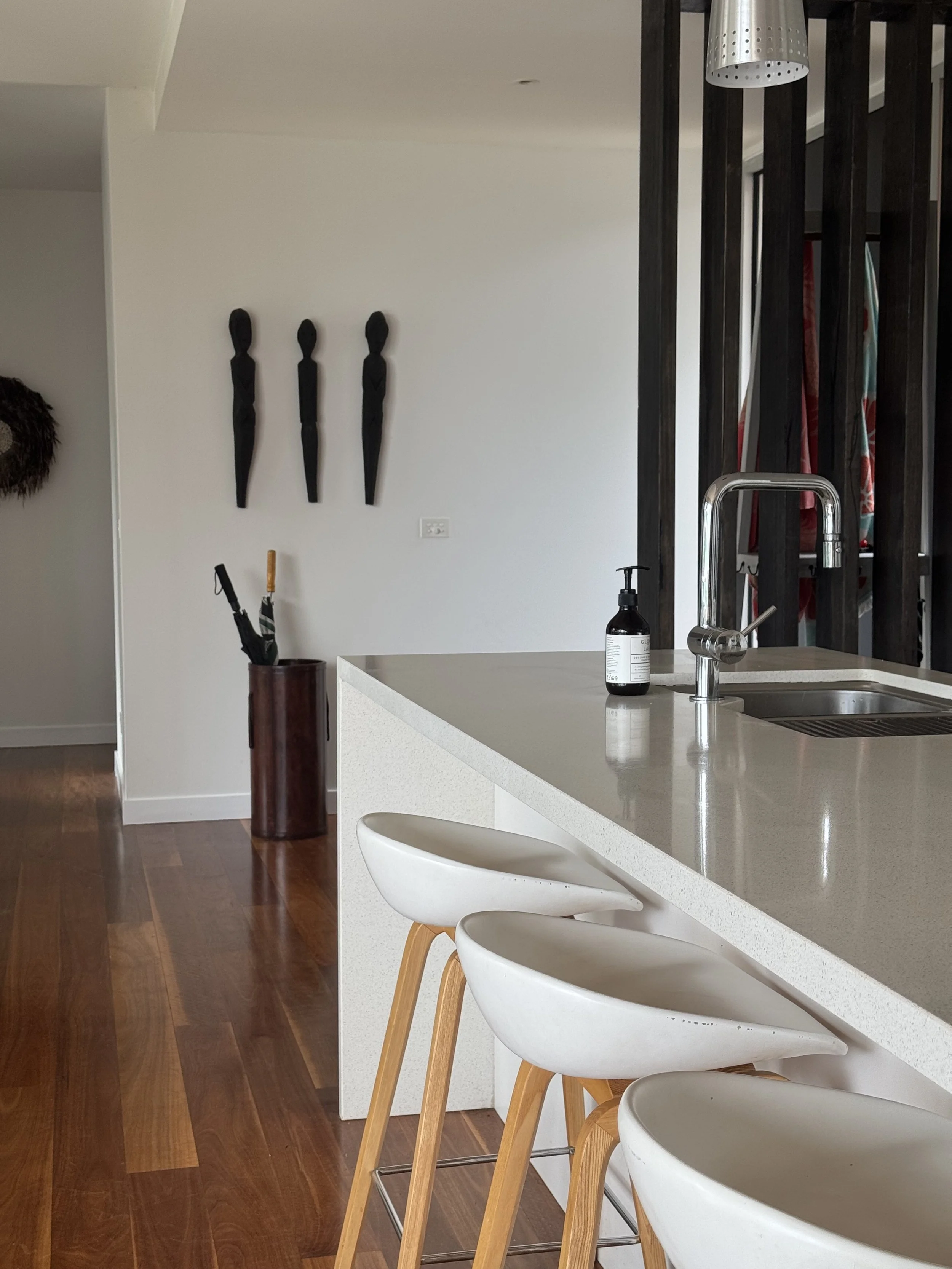 Modern kitchen with white countertop, three white and wooden bar stools, a black soap dispenser, and three black wall sculptures resembling abstract human figures.
