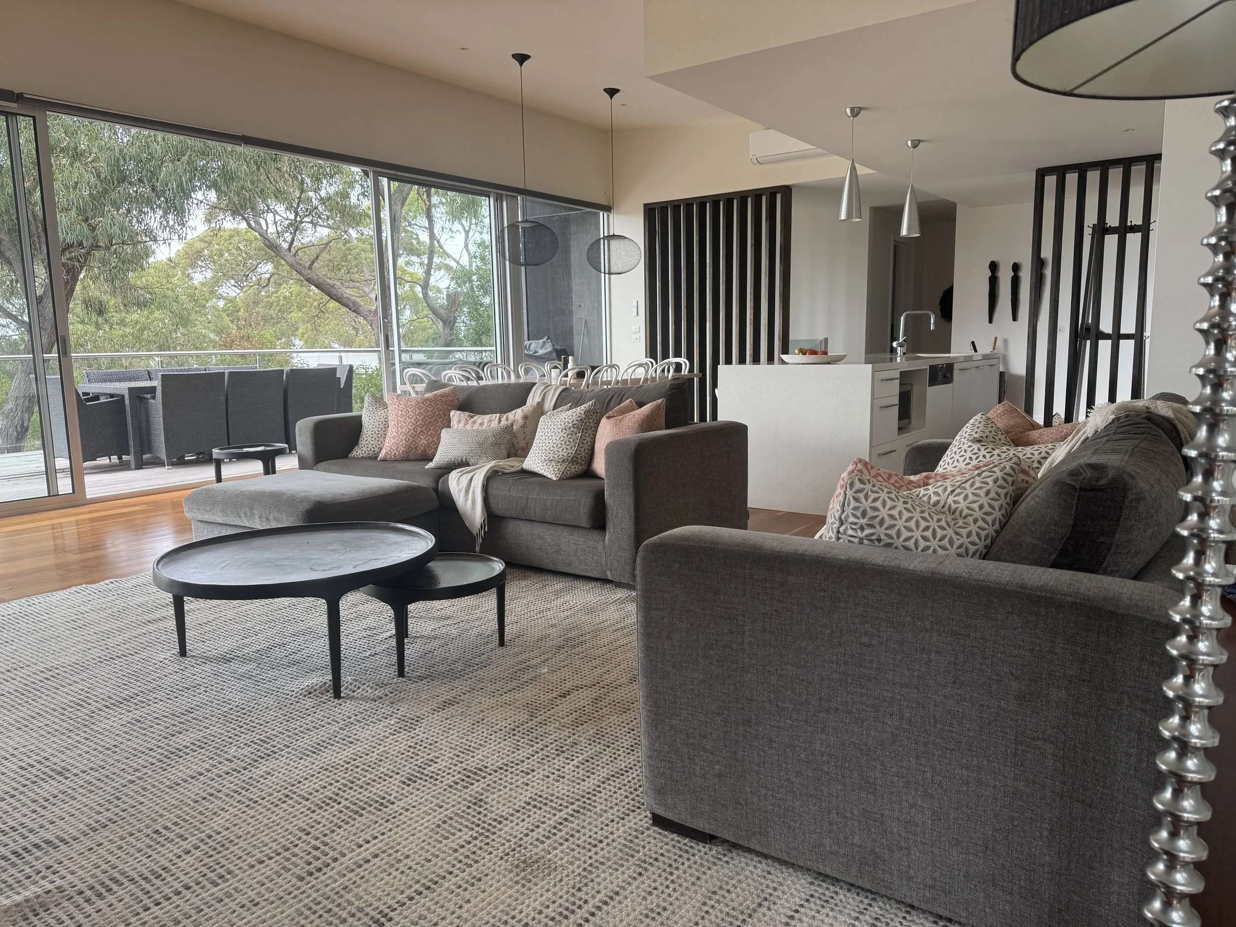 Living room with gray sofas, patterned pillows, black circular coffee tables, a large sliding glass door opening to an outdoor deck with trees, a modern kitchen with white cabinetry, and hanging pendant lights.