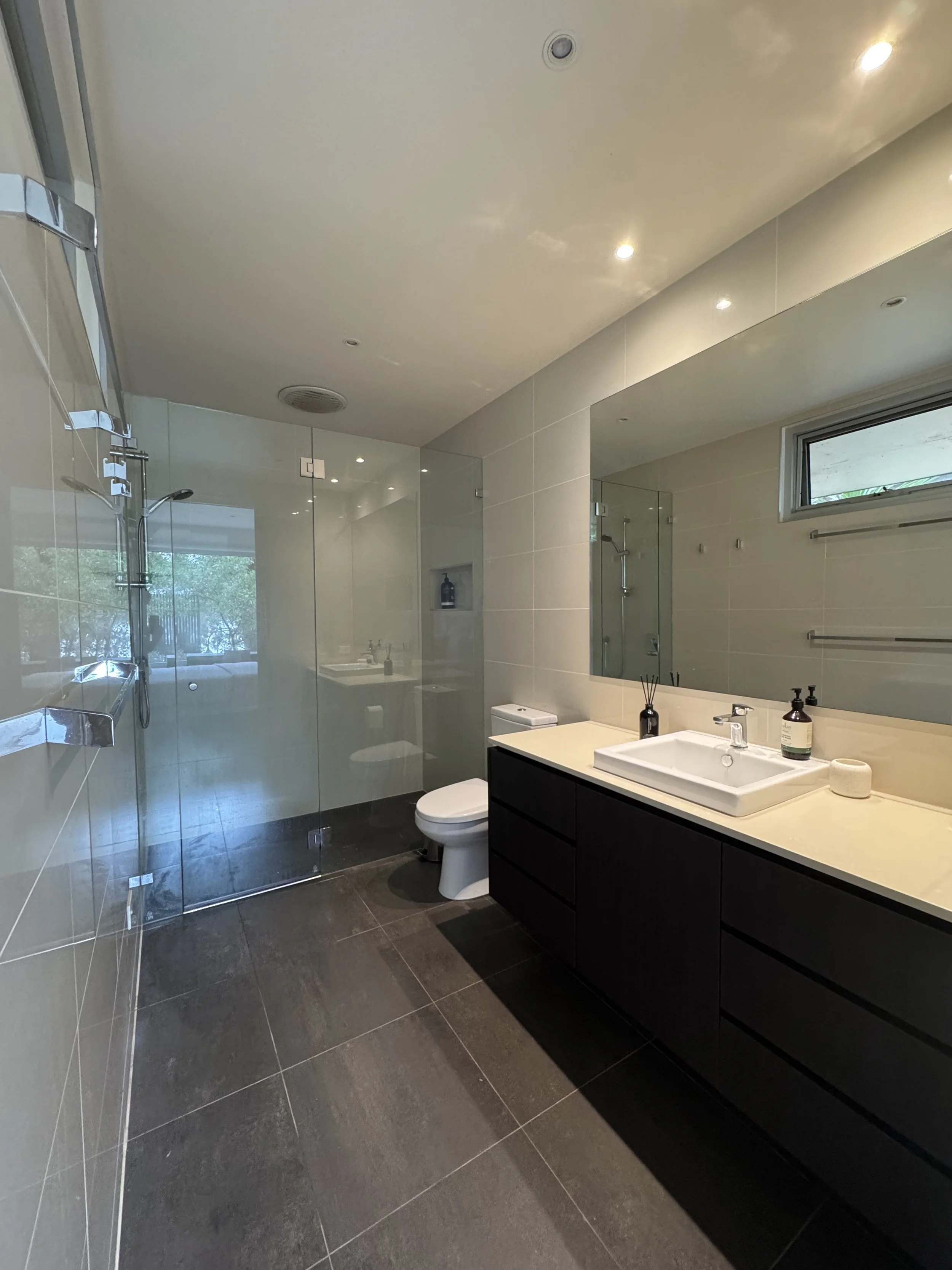 Modern bathroom with a glass-enclosed shower, dark tiled floor, white sink on a black cabinet, small window, and large mirror