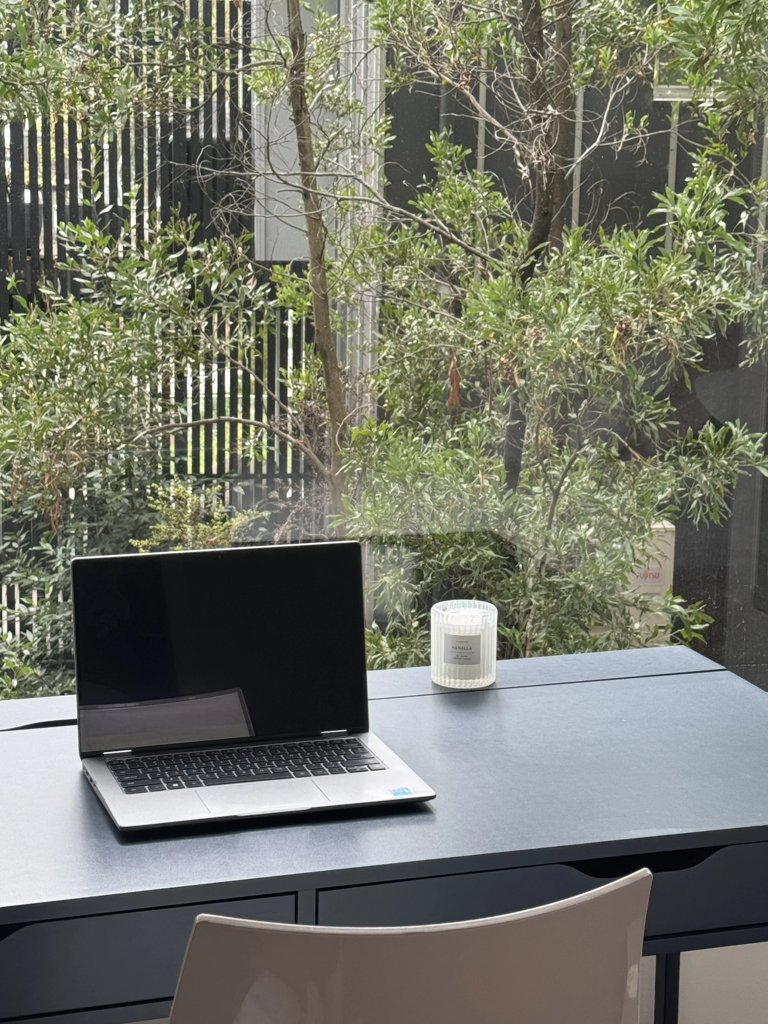 A laptop computer on a desk in front of a large window with a view of green trees and bushes outside. There is a white candle in a glass holder on the right side of the desk.