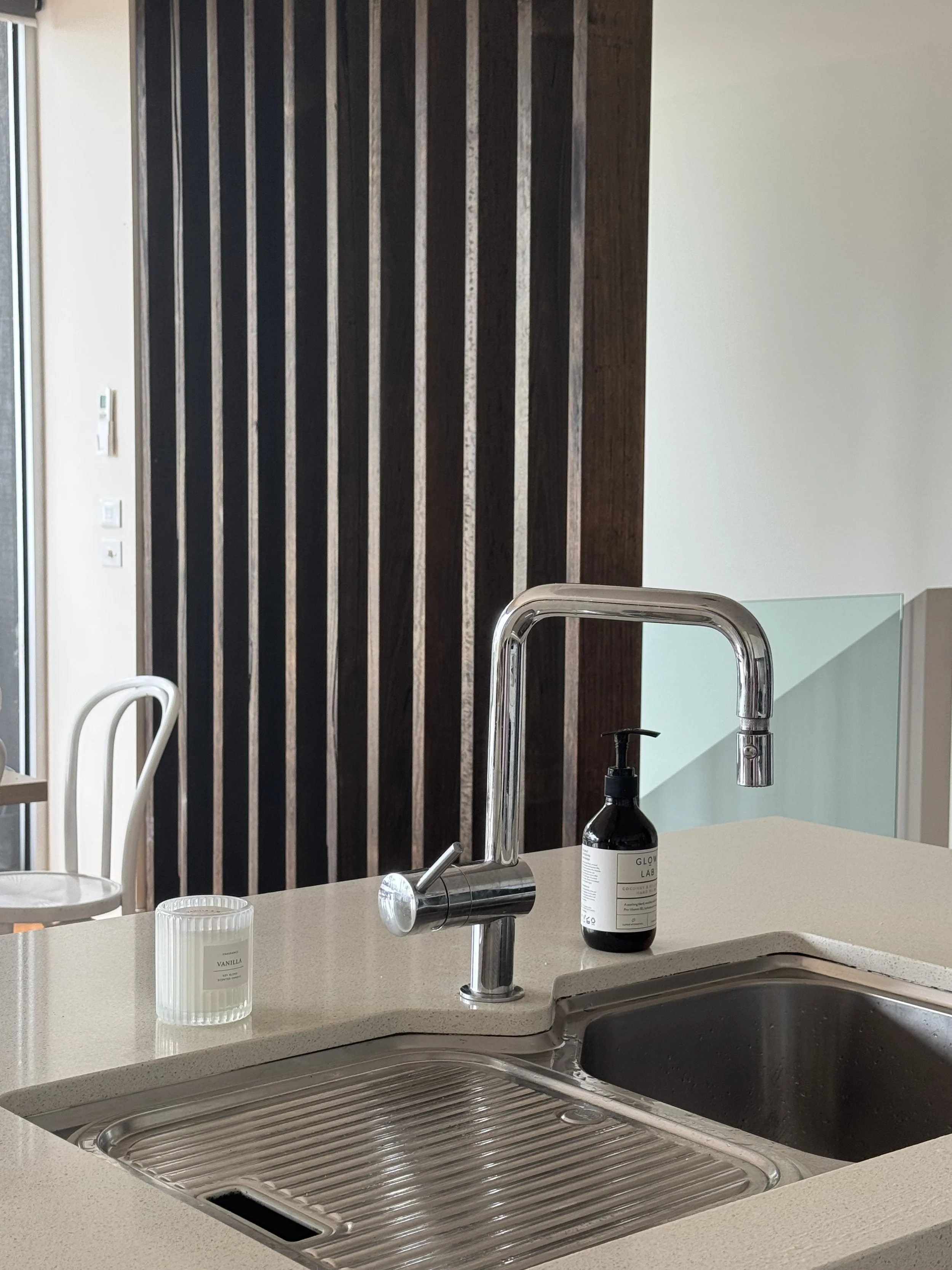 Close-up of kitchen sink area with a modern chrome faucet, a black soap dispenser, a small candle, and a double sink, with a dark wooden wall in the background.