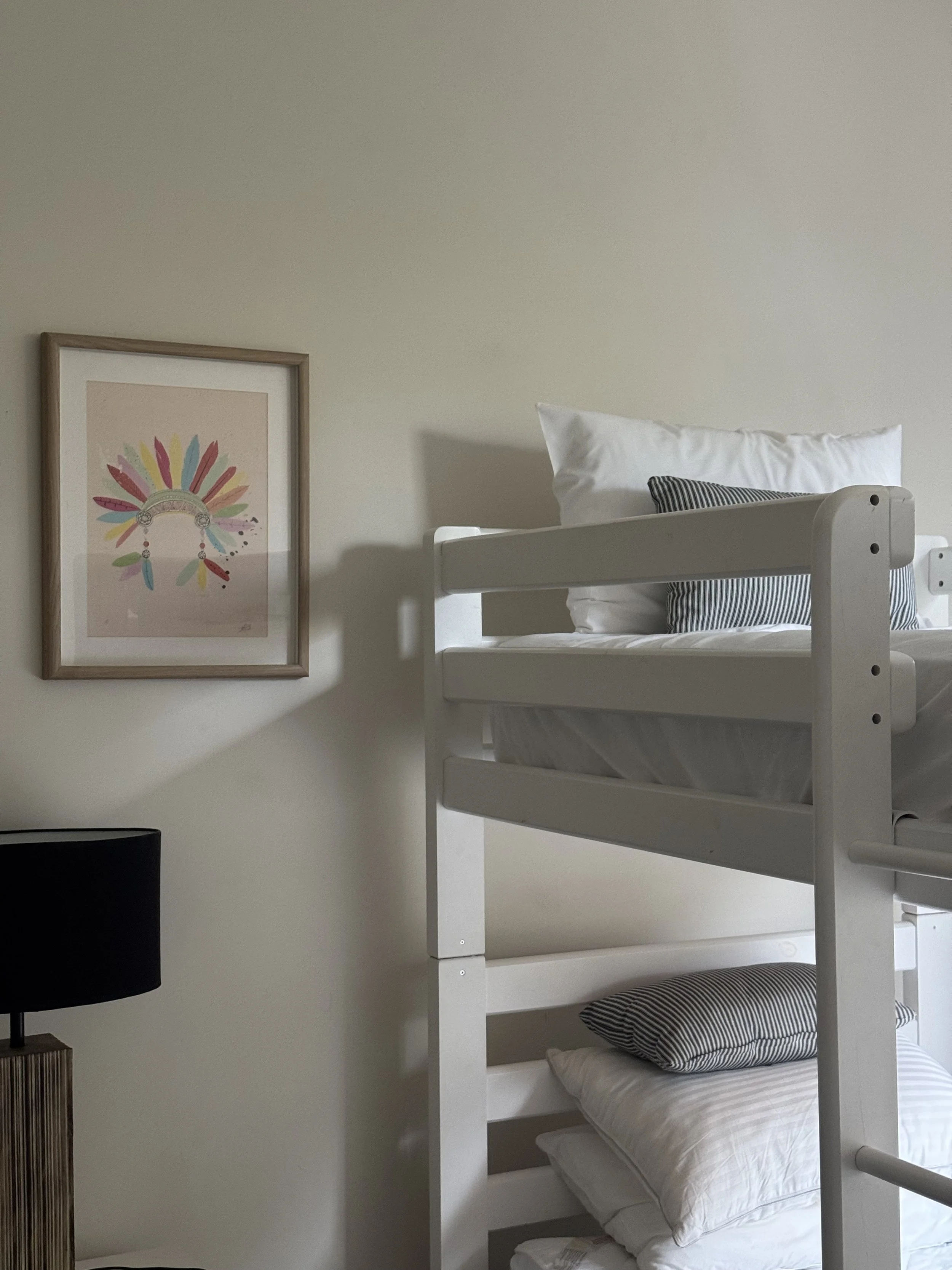 A bedroom with a white bunk bed with striped pillows, a framed colorful feather and arrow artwork on the wall, and a black lampshade on a wooden side table.