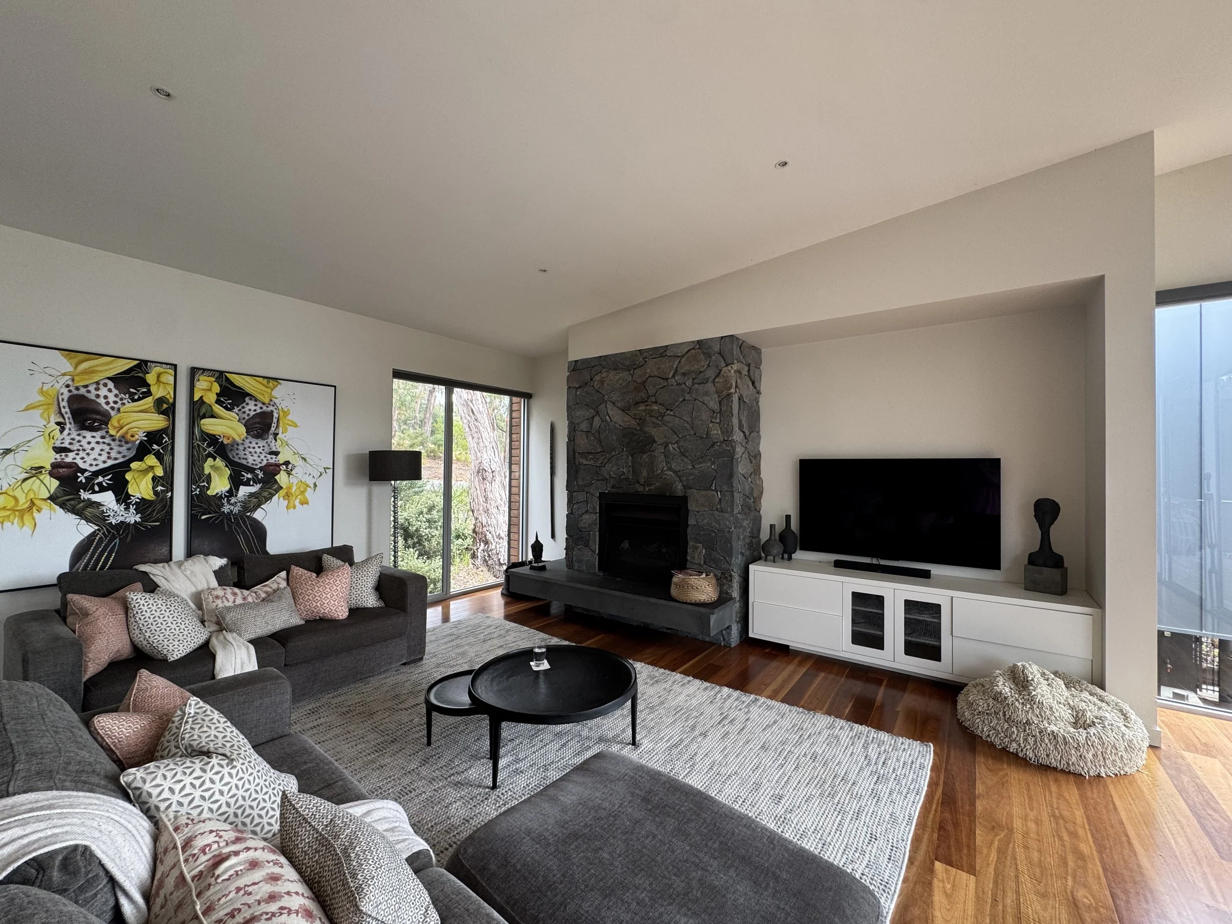 Living room with a dark gray sofa, patterned pillows, a large artwork of a woman's face with yellow flowers, a stone fireplace, a flat-screen TV, white console, and a floor-to-ceiling window showing trees outside.
