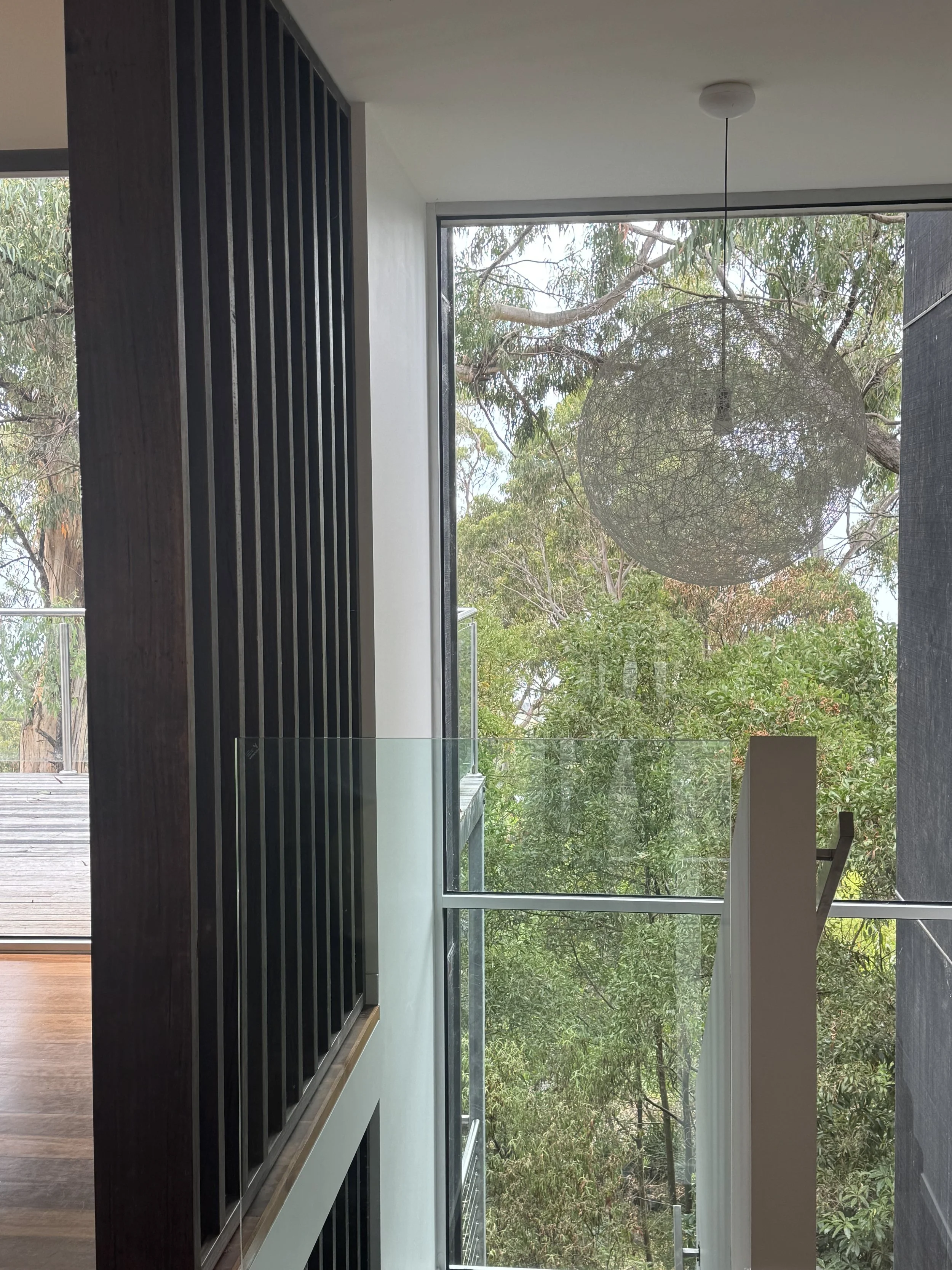 Interior view of a modern house featuring a large window with a view of trees outside, a hanging pendant light, and a glass railing. Part of a dark wooden staircase and wall with vertical slats are visible.