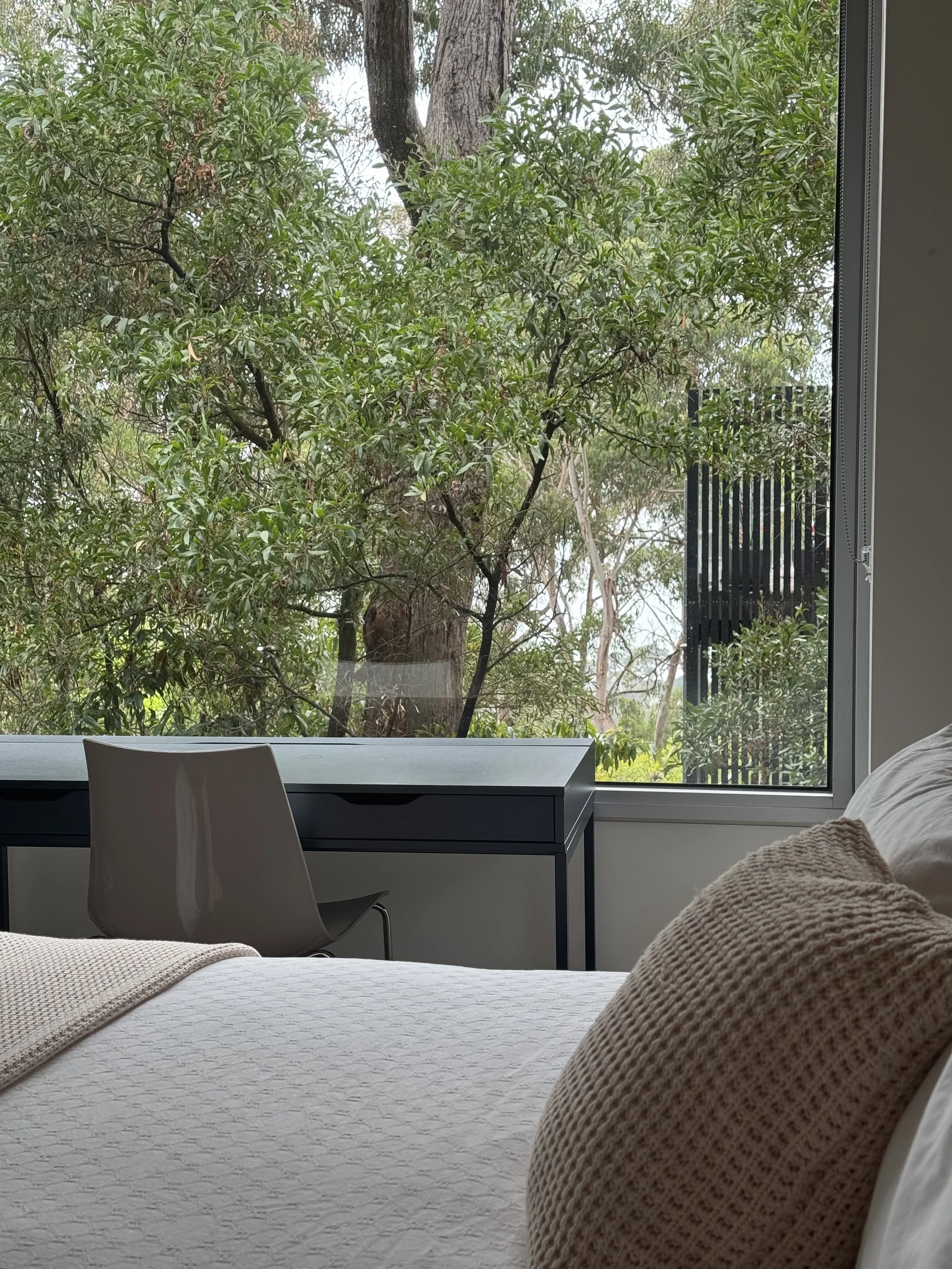 A bedroom with a bed covered in a light-colored quilted blanket and beige textured pillows, next to a window with greenery outside, including trees and bushes, and a black outdoor structure visible through the window.