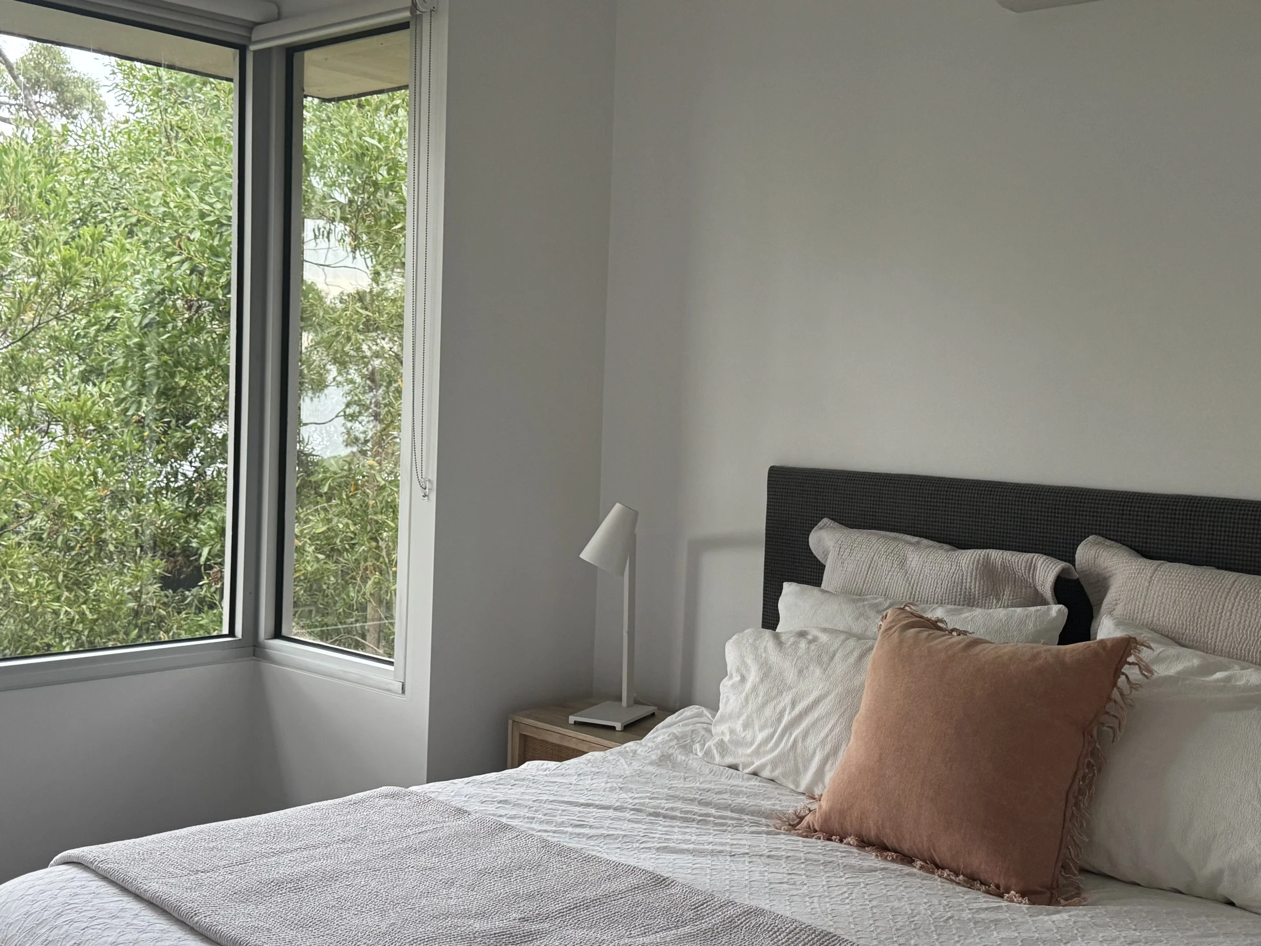 A bedroom with a bed, white bedding, multiple pillows, and a peach-colored pillow. There is a wooden nightstand with a white desk lamp, and a large window showing green trees outside.