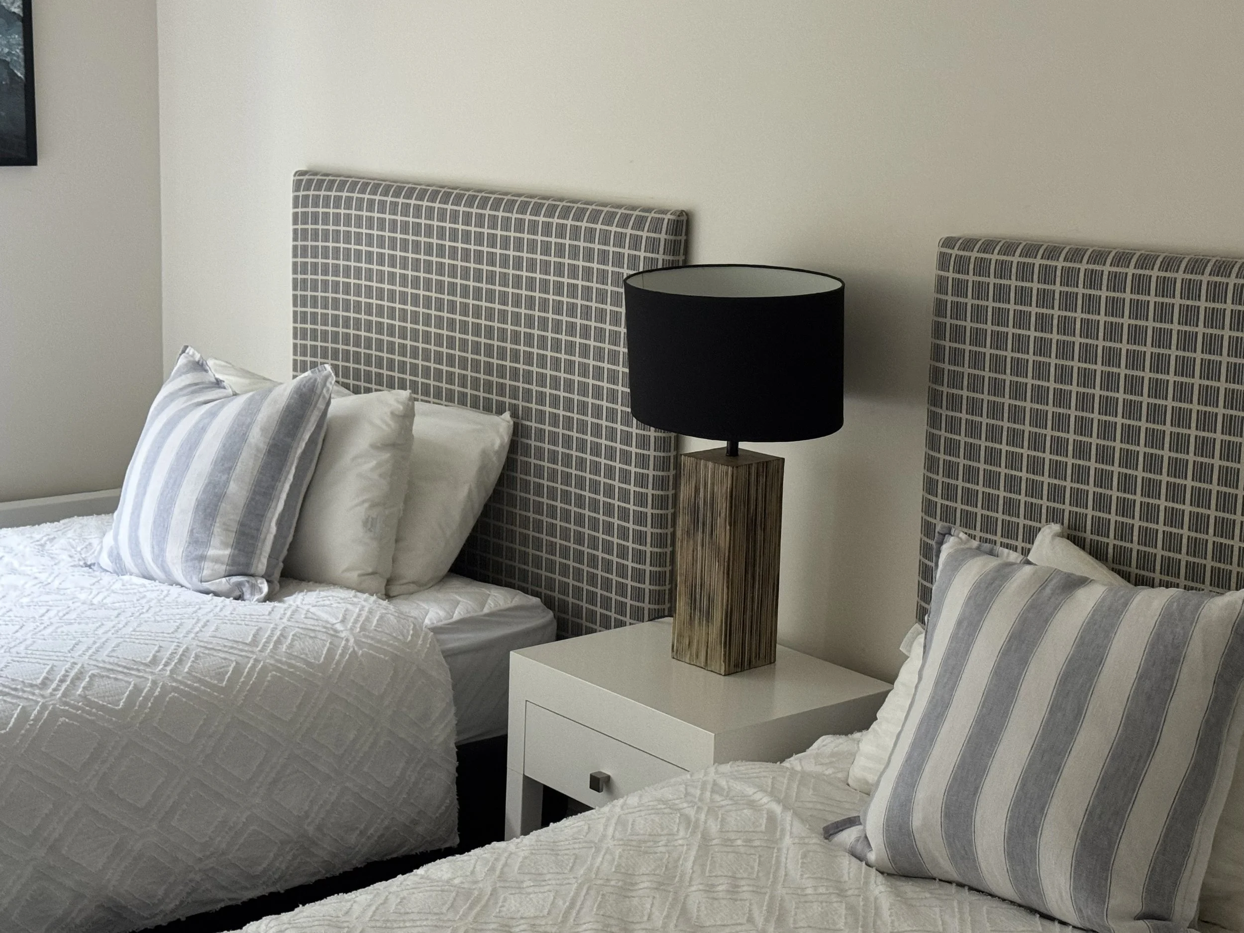 Two beds with patterned headboards, each with pillows and white textured bedspreads, and a white nightstand with a wooden table lamp with a black shade.