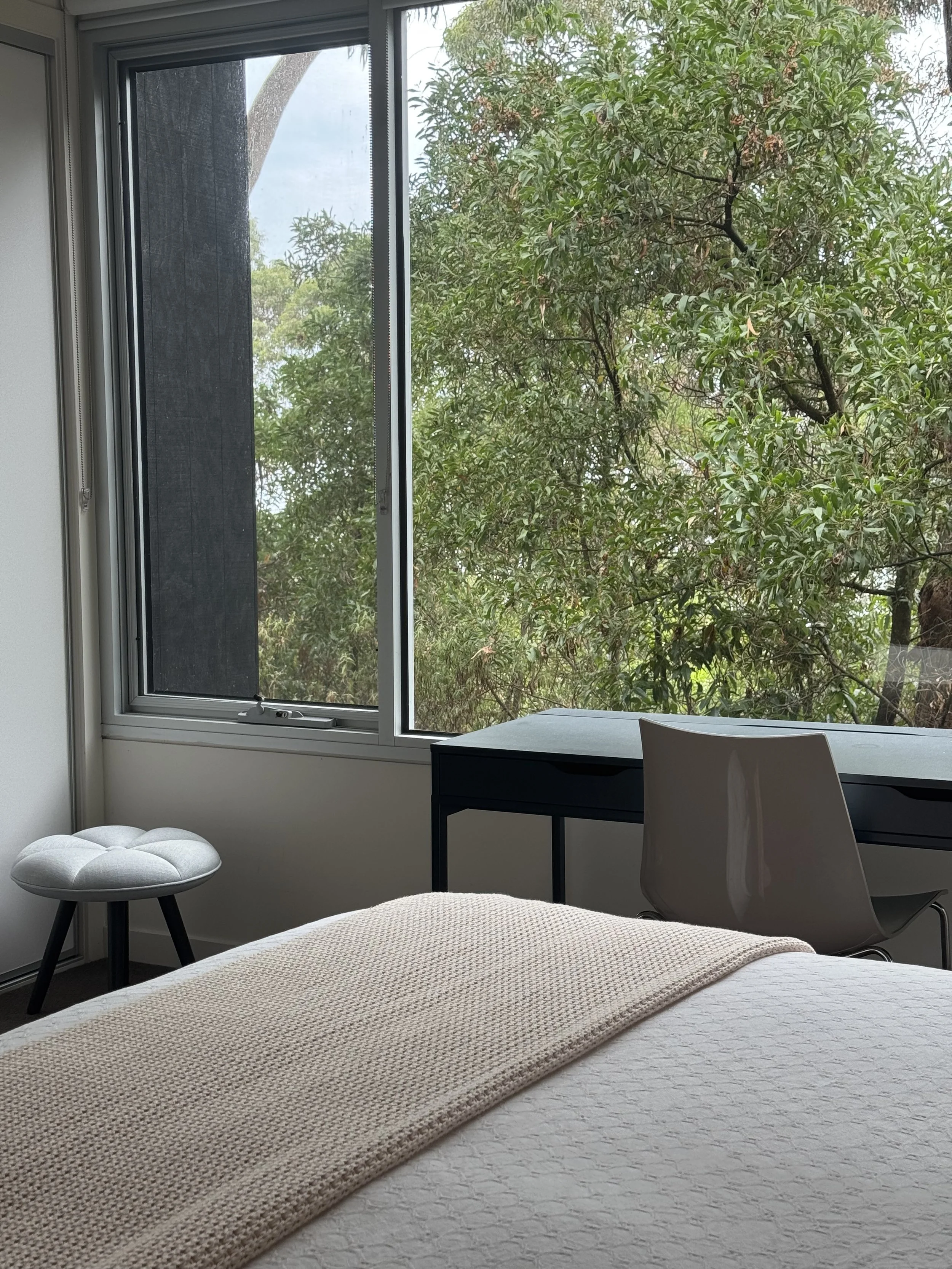 Bedroom with a bed covered by a beige blanket, a small gray stool, a desk with a gray chair, and a large window showing green trees outside.