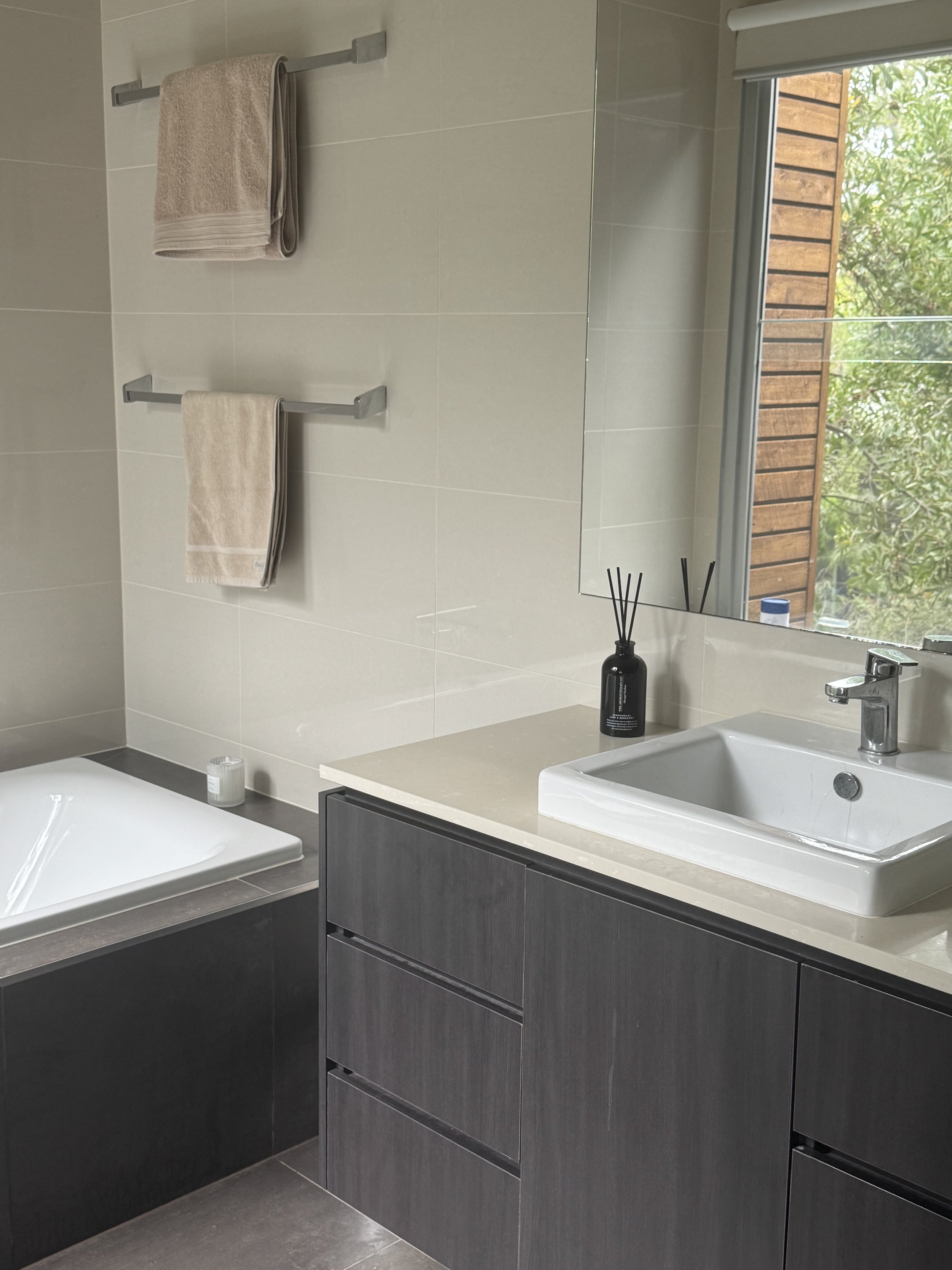 Modern bathroom with a dark gray vanity, white sink, black diffuser, beige towels on a towel rack, window with a view of green trees, and a bathtub.