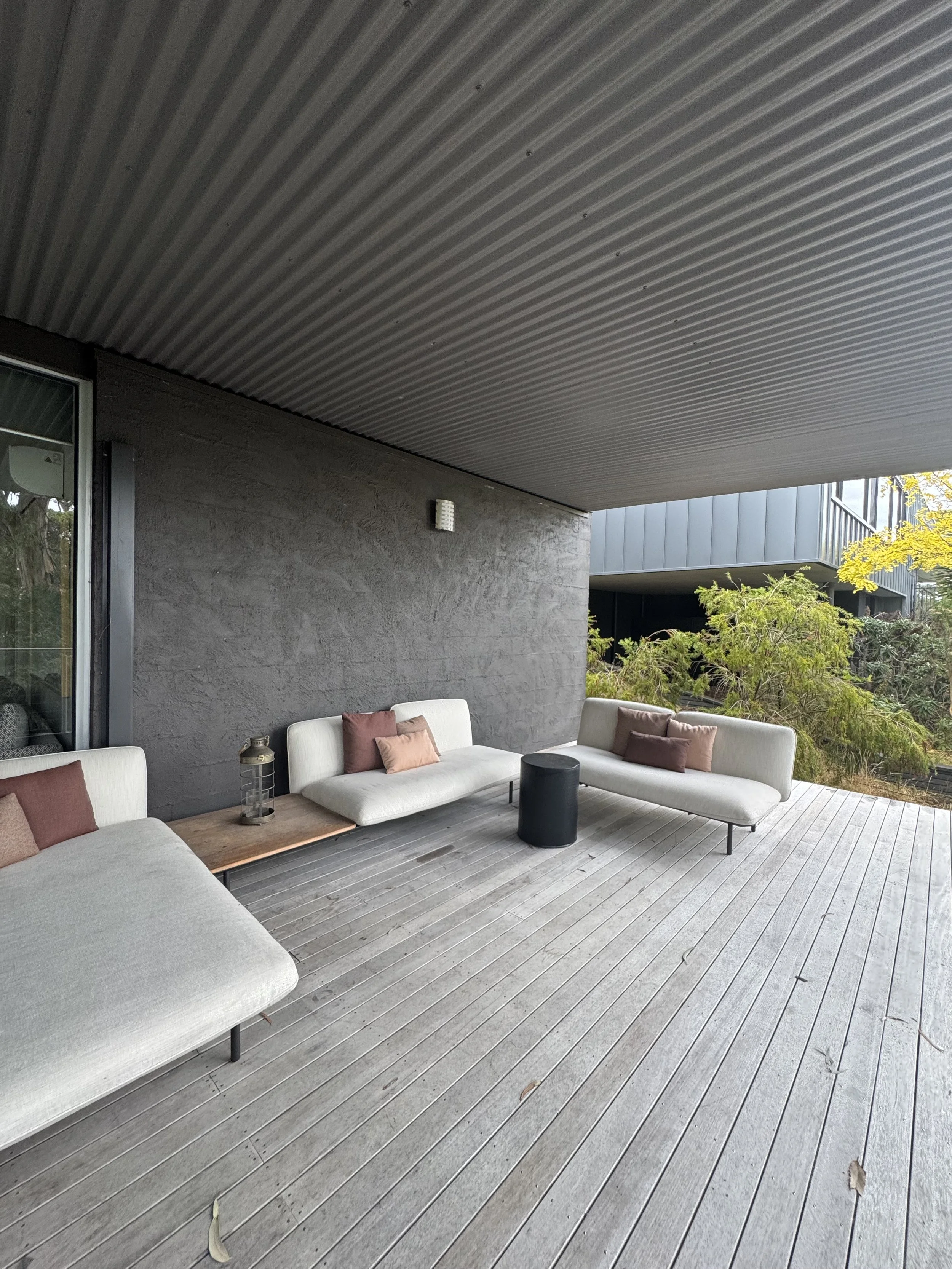 A modern outdoor patio with three white cushioned sofas, each decorated with pink and brown pillows. A small black round table is placed between two sofas, with a lantern on a wooden side table next to one of the sofas. The patio has wooden flooring 