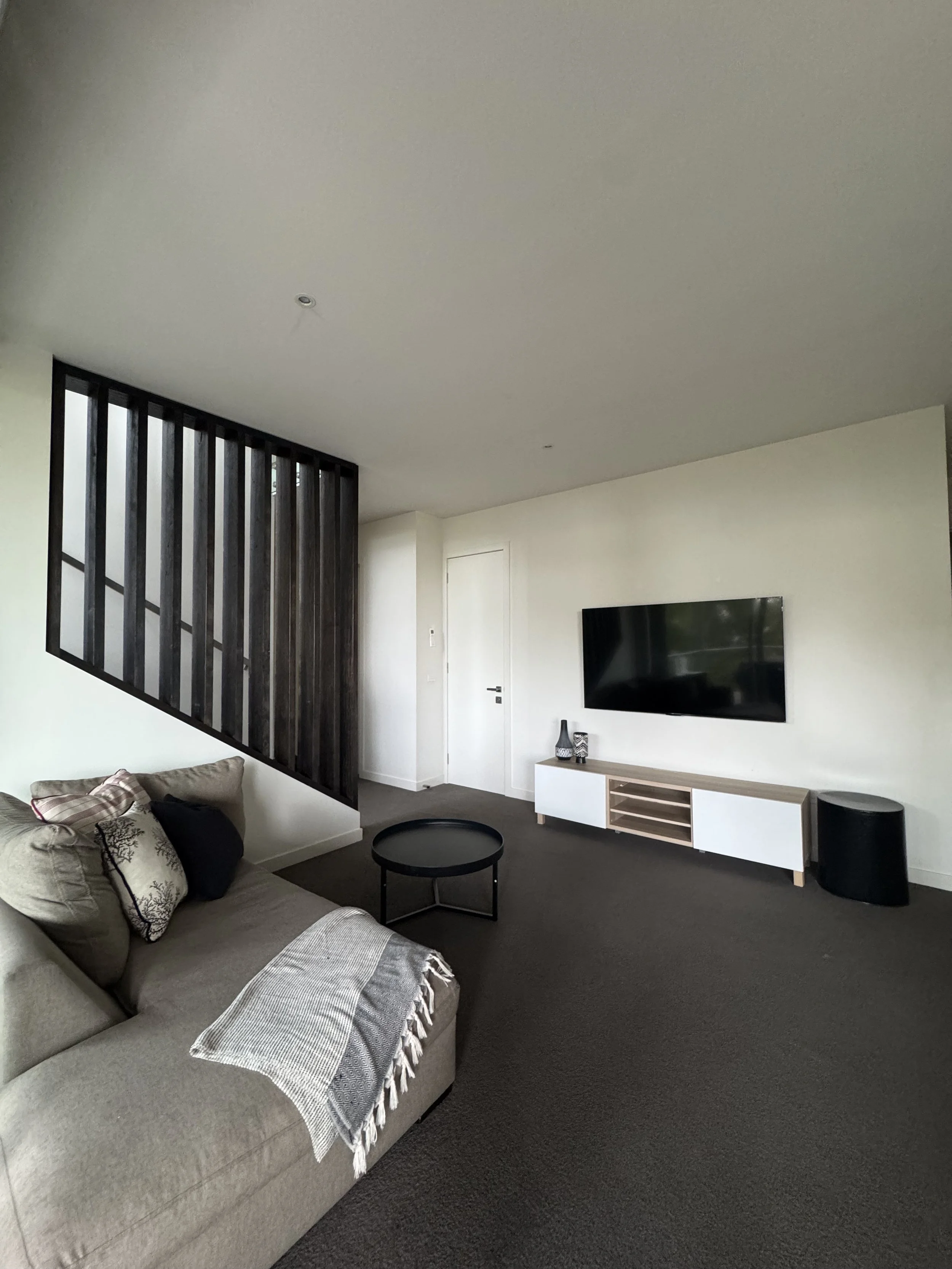 Living room with beige sofa, throw pillows, black coffee table, wall-mounted TV, wooden media console, black side table, dark carpet, wooden staircase with black railing, and white walls.