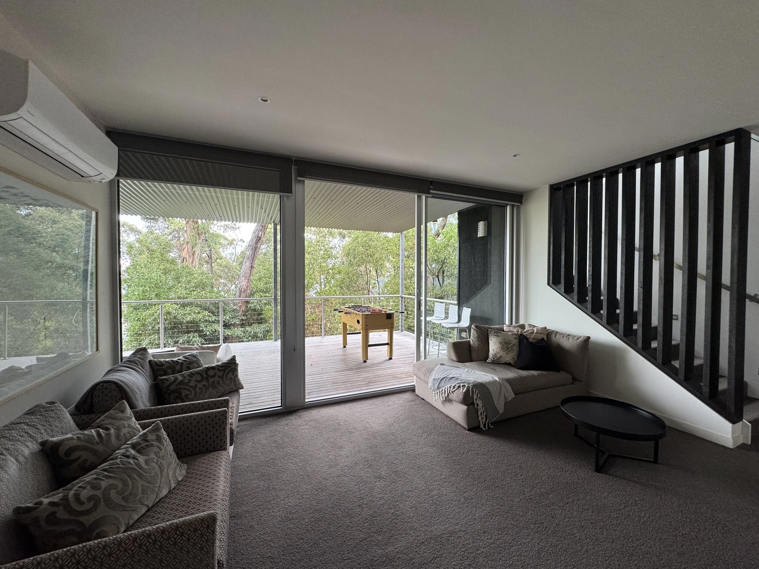Living room with a sliding glass door leading to a balcony and trees outside, a beige sofa with patterned cushions on the left, a cream-colored couch with throw pillows and a blanket on the right, a small black coffee table, and a foosball table on t