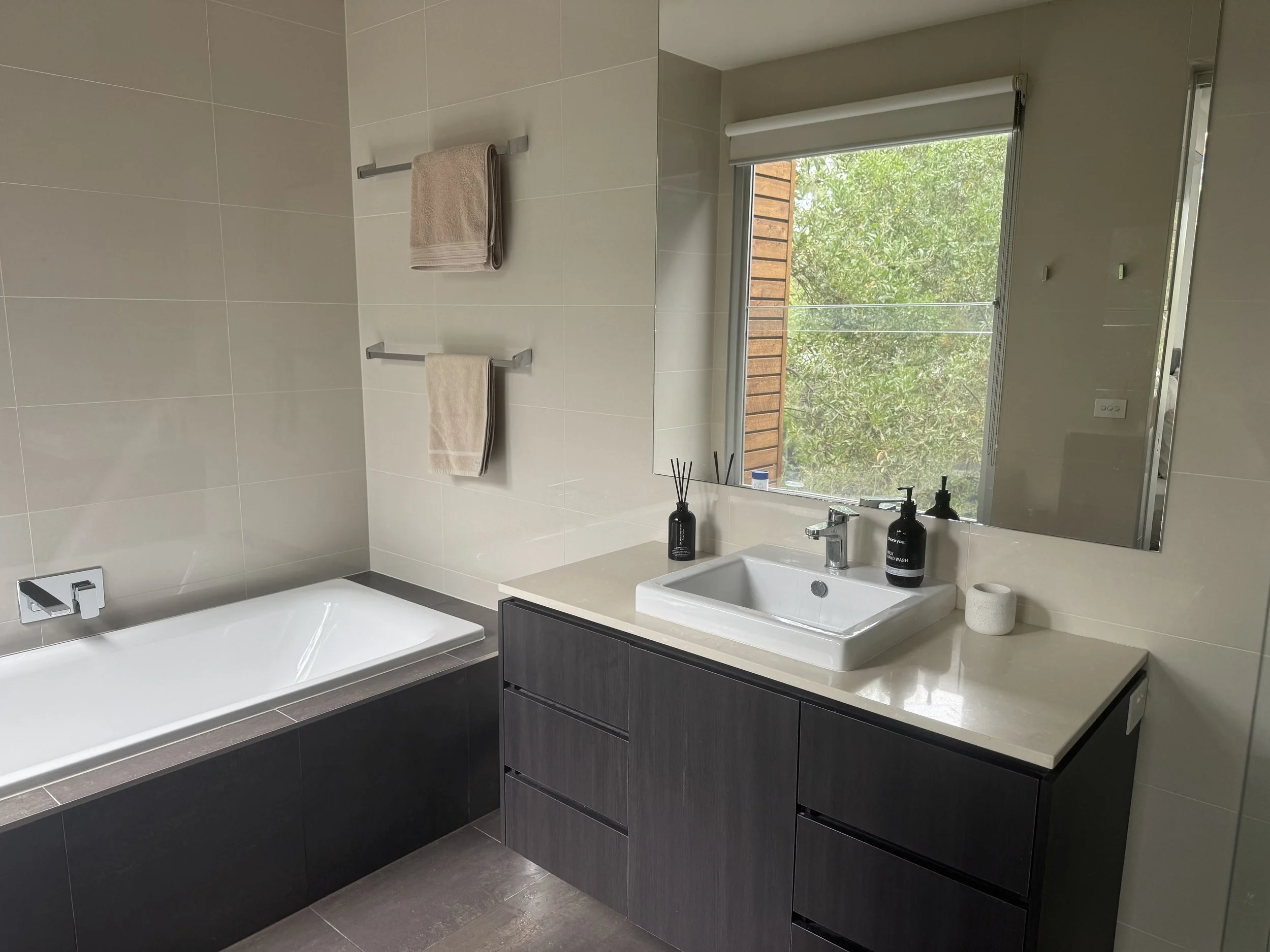 Modern bathroom with a bathtub on the left, a vanity with a rectangular sink, soap dispenser, and a reed diffuser, a large mirror, window showing greenery outside, beige towels on wall hooks, and neutral-colored tiles.
