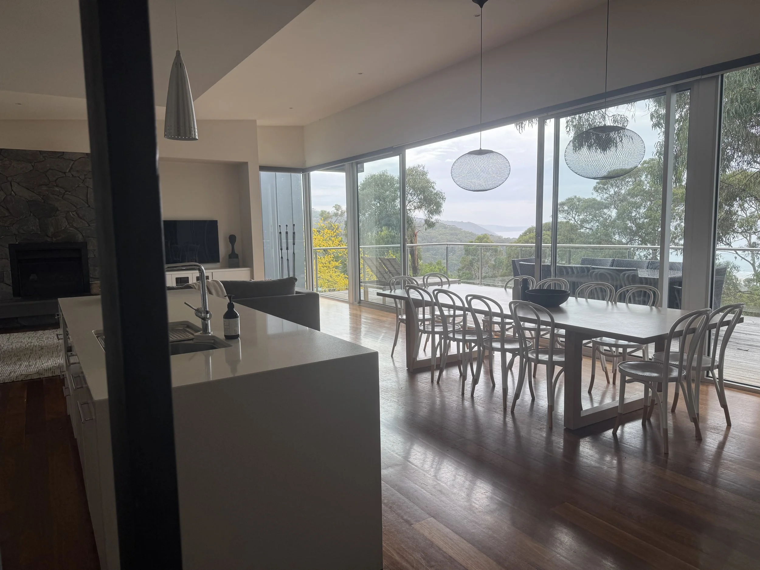 Open-concept living and dining area with a large wooden dining table surrounded by white chairs, large windows with a mountain and tree view, modern lighting fixtures, and a kitchen island with a sink and bottle, in a contemporary home.