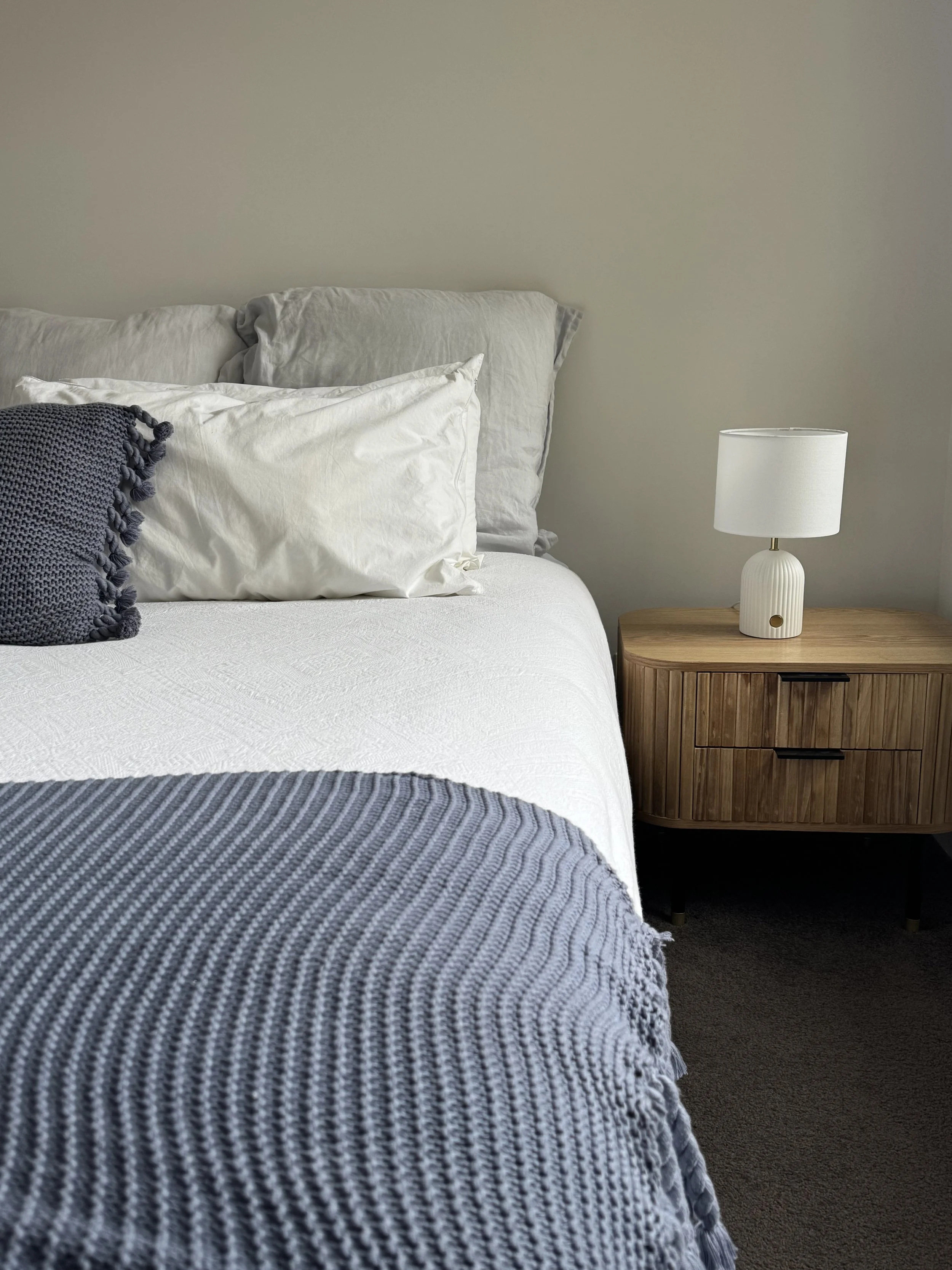 A neatly made bed with white and gray pillows and a textured gray blanket at the foot, next to a wooden nightstand with a white table lamp and a textured beige wall.