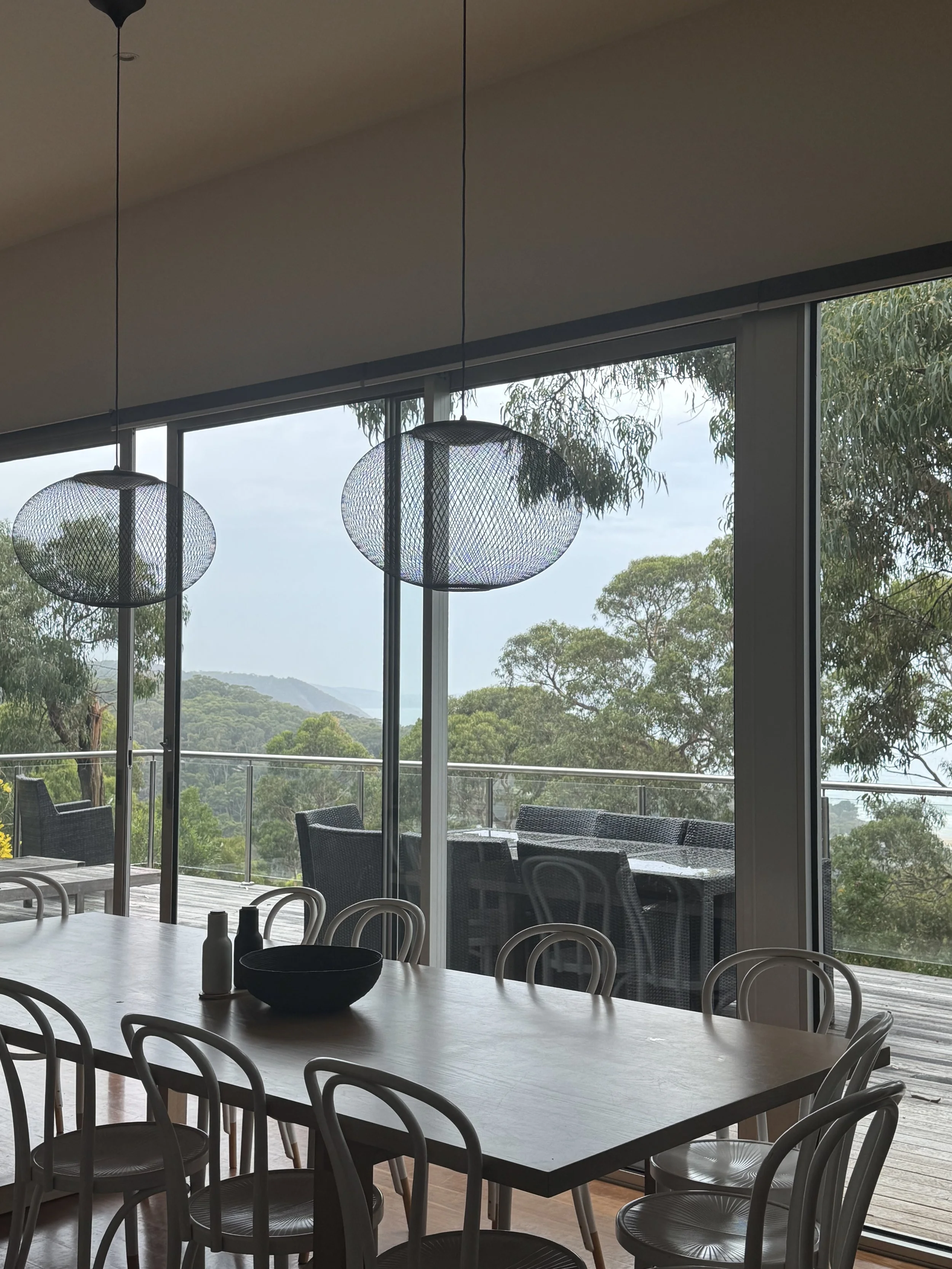 Interior of a dining area with a large wooden table, white chairs, and a view of a scenic outdoor deck with patio furniture and lush greenery through large sliding glass doors.