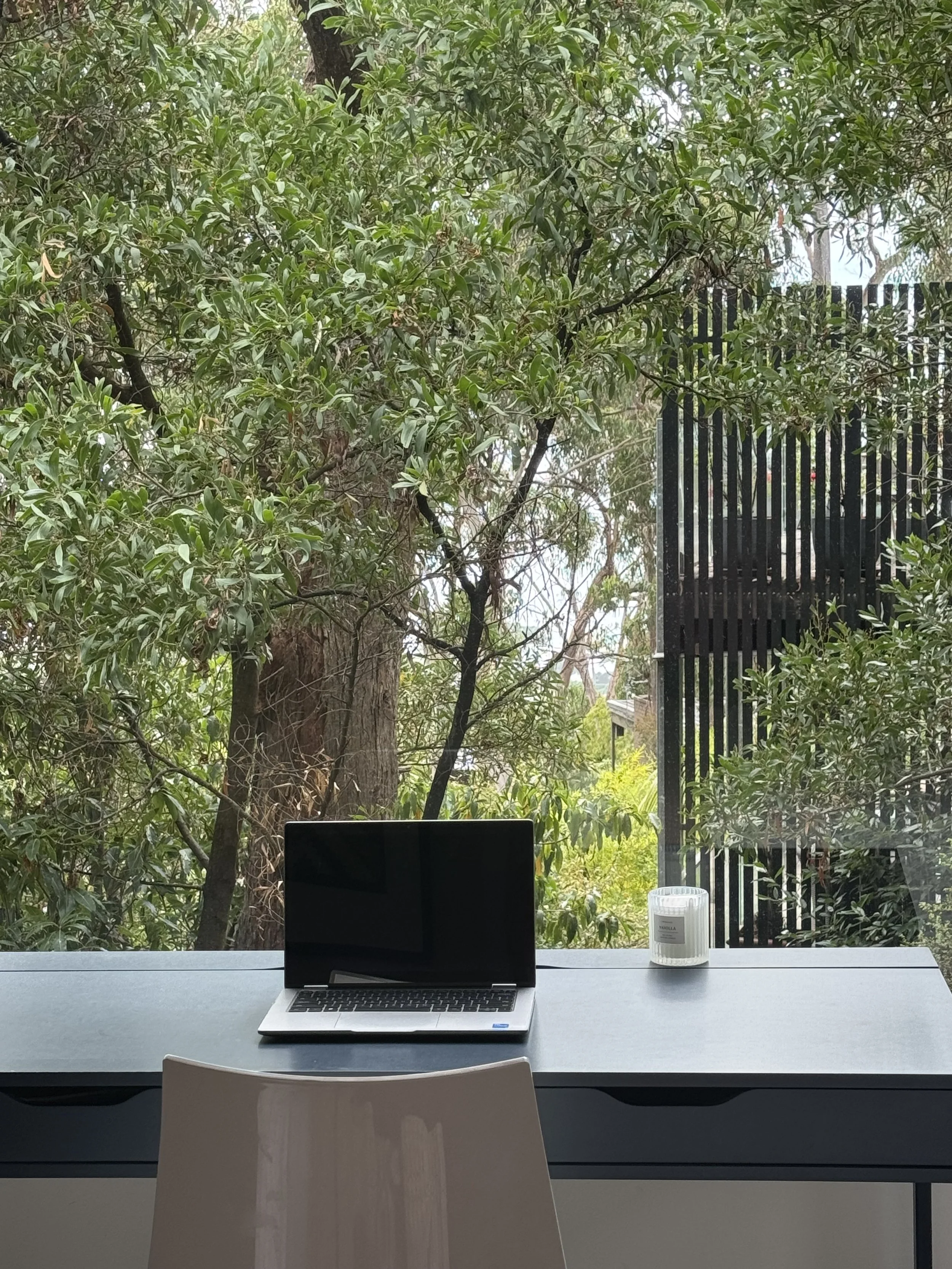 A work desk with a closed laptop and a paper candle holder, set against a large window showing trees and greenery outside.