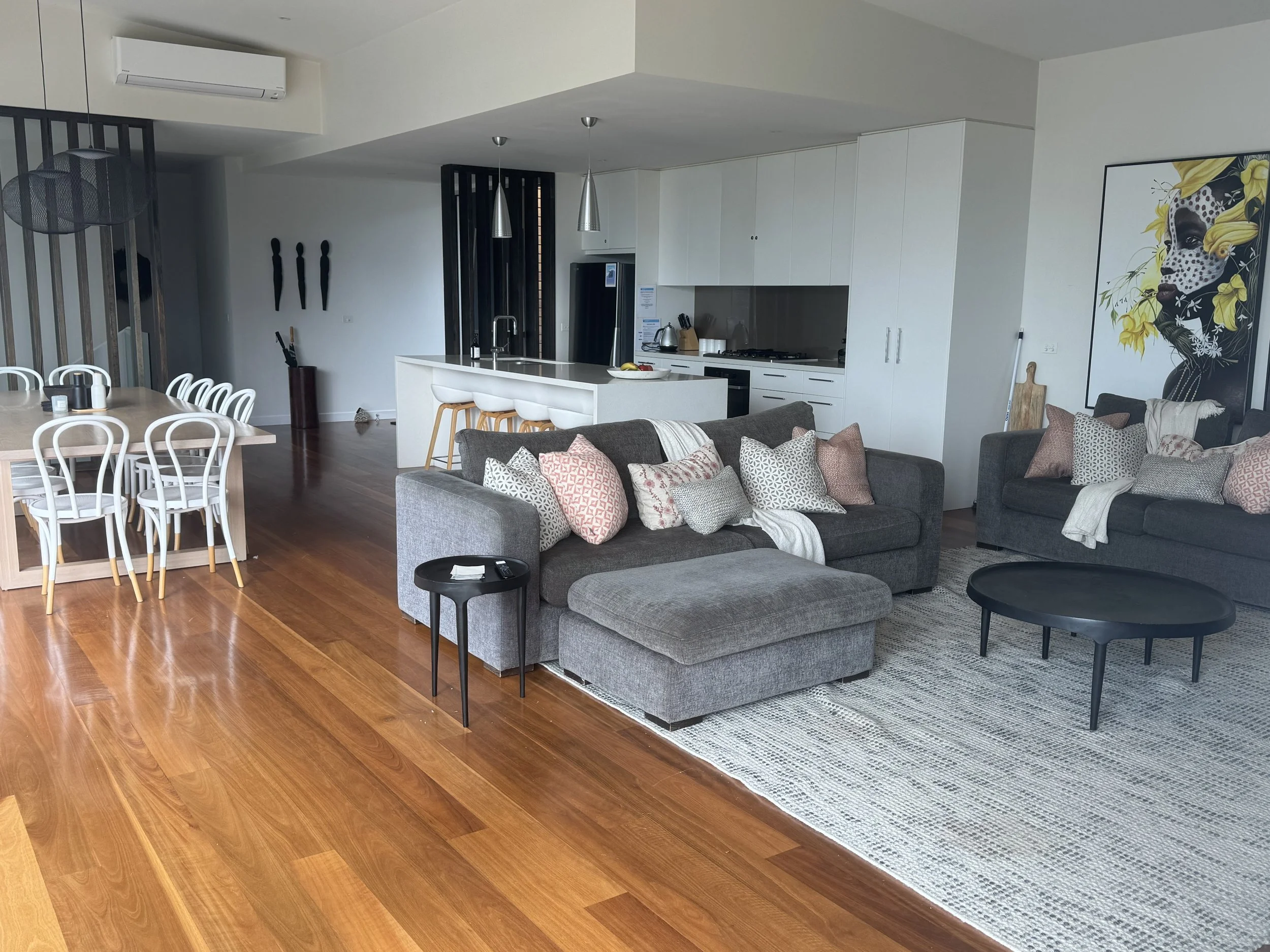 Open plan living space with a gray sectional sofa, a black oval coffee table, and a large wall art of a woman with yellow flowers on her head. The area includes a kitchen with white cabinets, a white island with bar stools, and a dining table with wh