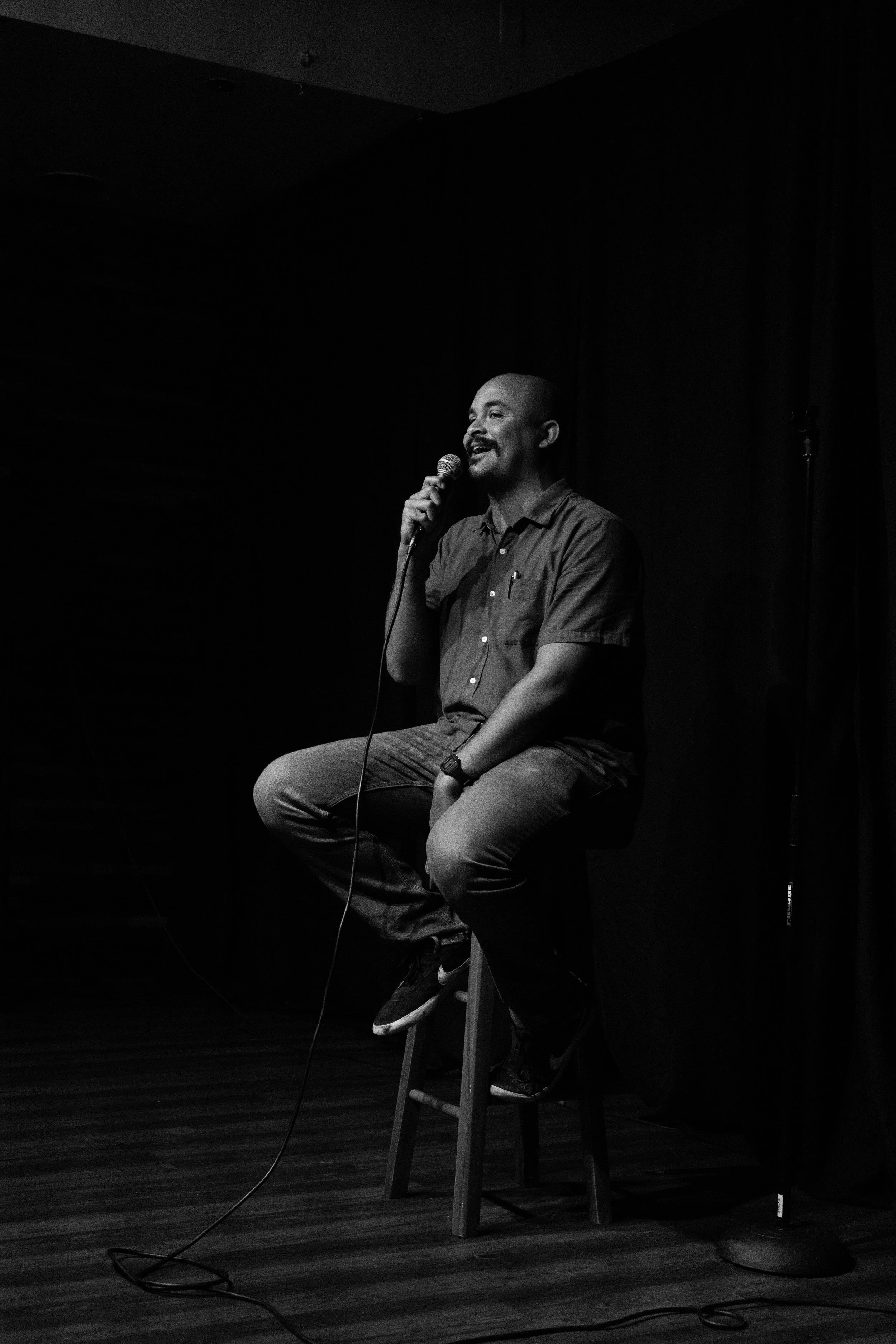 A man sitting on a stool on a stage, holding a microphone and performing stand-up comedy in a dark room.