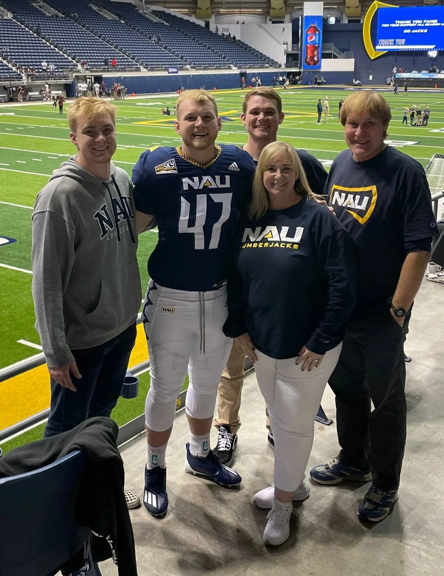 Group of four people standing on the sidelines of a football field in a stadium, smiling at the camera. The person second from the left is wearing a football uniform with the number 47 and NAU (Northern Arizona University) logo. The others are dressed casually, and the stadium sits mostly empty with some spectators visible in the background.