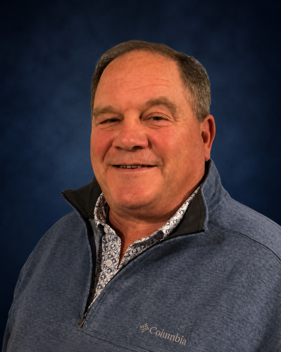 A smiling middle-aged man with short brown hair wearing a Columbia fleece jacket and a patterned shirt, standing against a dark blue background.