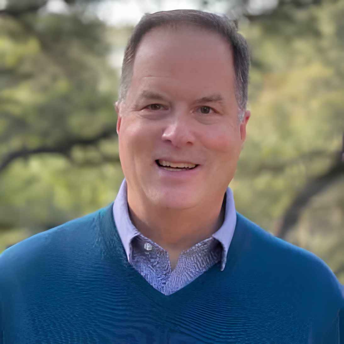 Portrait of a man smiling outdoors with blurred trees in the background.
