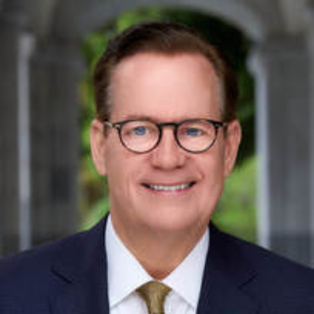 Professional headshot of a middle-aged man with glasses, brown hair, wearing a dark suit, white shirt, and a light-colored tie, outdoors with blurred greenery in the background.