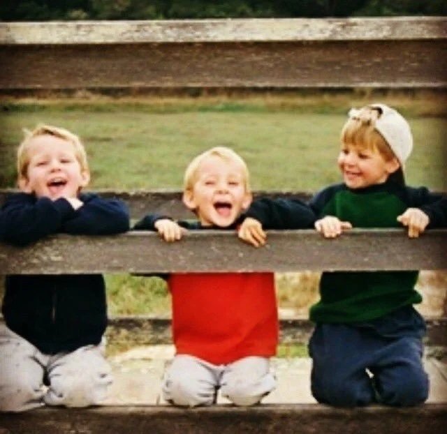 Three children laughing and playing behind a wooden fence outdoors.