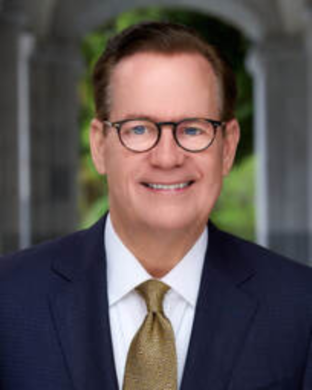 A professional headshot of a man in a dark suit, white shirt, and gold tie, with glasses, smiling outdoors.