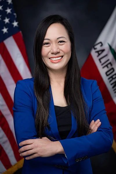 A woman with long black hair wearing a blue blazer, smiling, with American and California flags in the background.