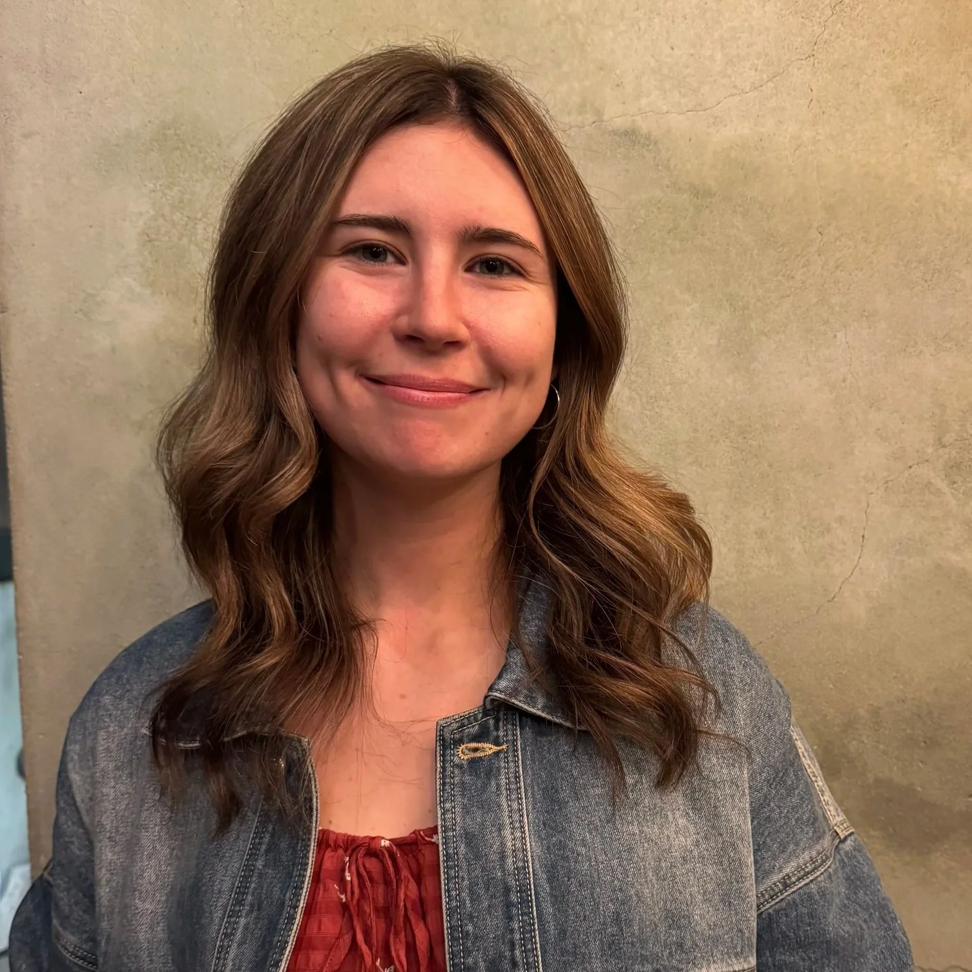 Young woman with wavy brown hair smiling against a beige textured wall, wearing a denim jacket and red top.