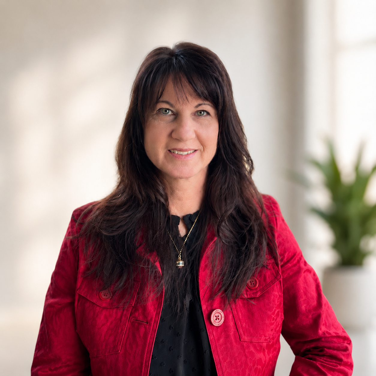 A woman with long, wavy dark hair and light skin, smiling, wearing a red jacket over a black top, with a gold necklace, standing indoors with a blurred background and a green plant.
