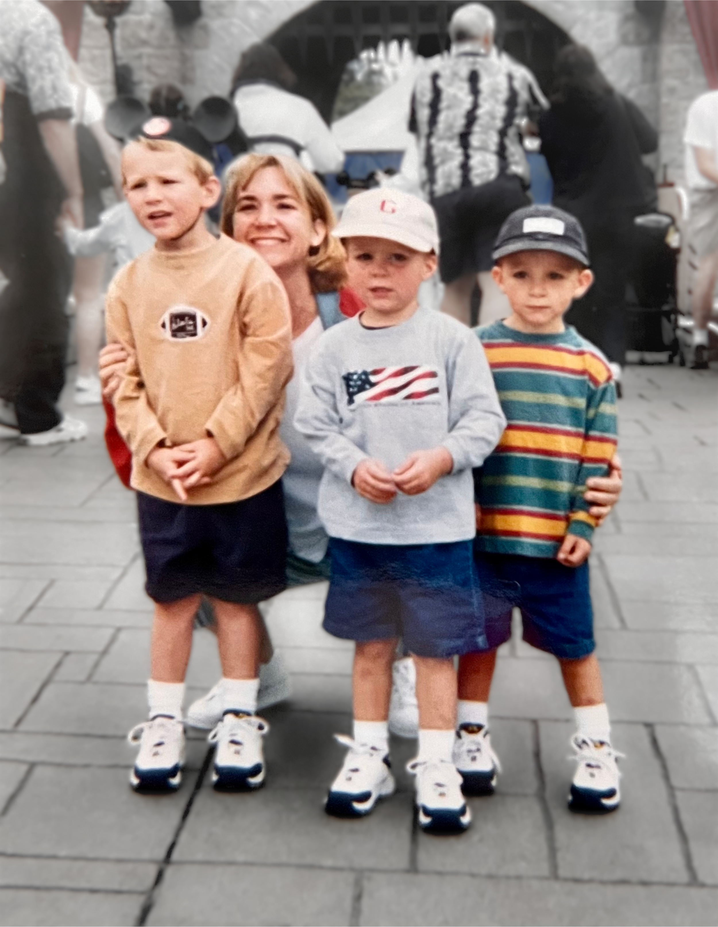 A woman with three young boys posing in front of a castle-like structure, all wearing sneakers, shorts, and casual clothing. The boys are standing with their arms crossed or by their sides, and the woman is smiling.