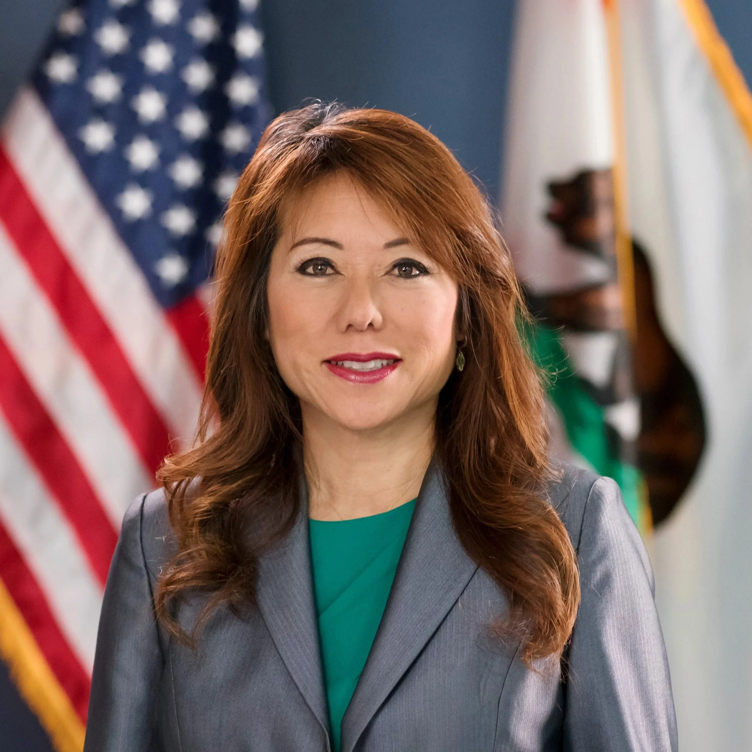 A woman with reddish-brown hair and wearing a gray blazer and green top, standing in front of a blurred background with American flags and flags from other countries.
